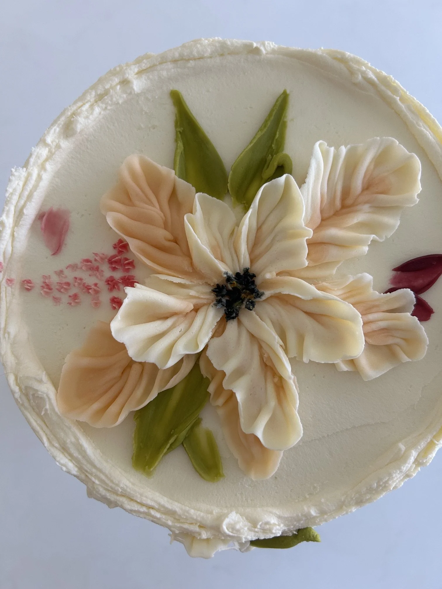 Close-up of a decorated cake featuring a large flower made of icing, with green leaves, pink accents, and small black details on a white cake surface.