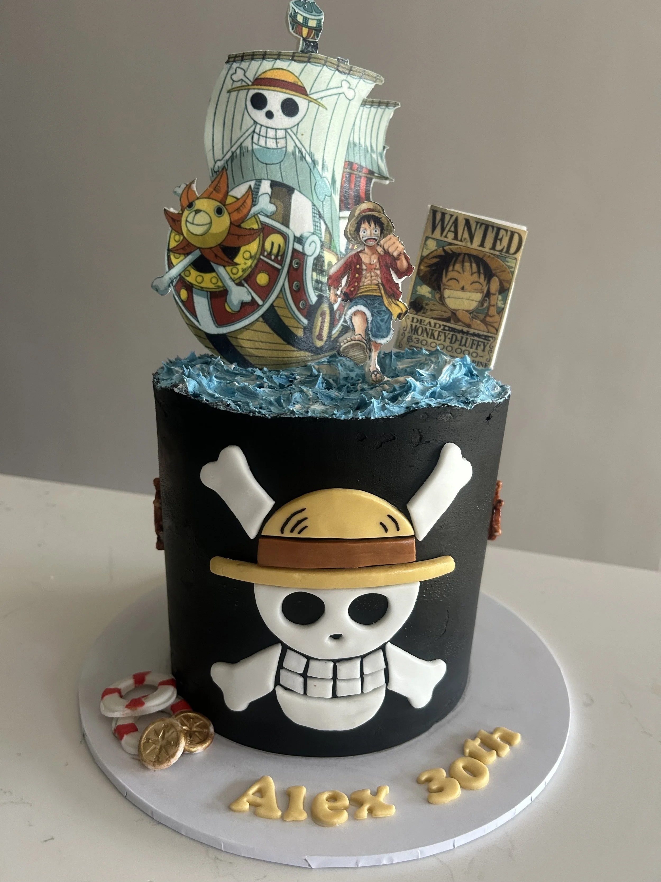 A Pirate-themed cake with a black skull and crossbones design, decorated with a skull and crossbones figure, a pirate ship, and characters from One Piece, including Monkey D. Luffy and Tony Tony Chopper, with gold coins, candy canes, and a "Alex 30" 