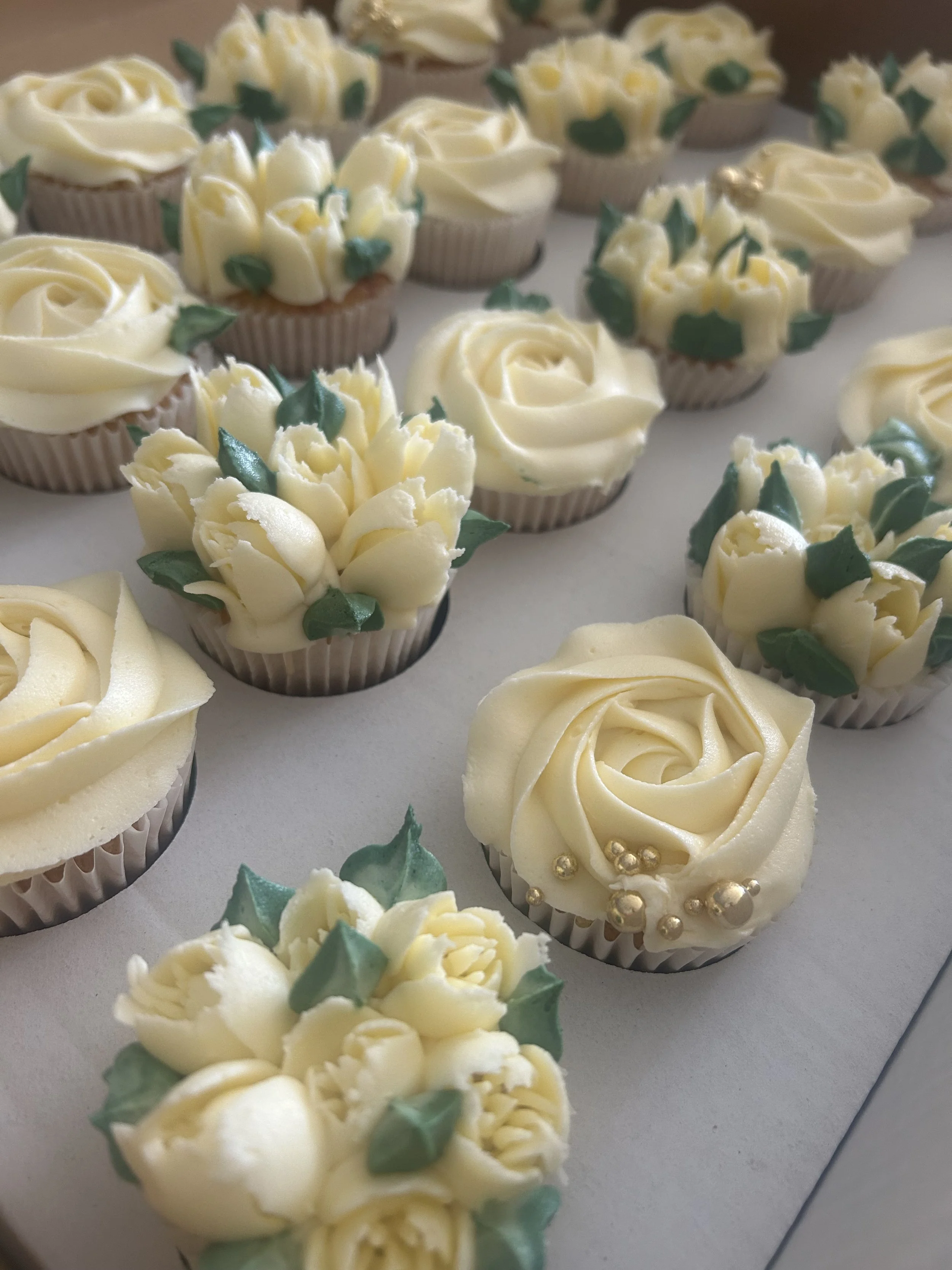 Assorted cupcakes decorated with white buttercream in rose and flower shapes, some with green leaf accents and gold-colored edible beads.