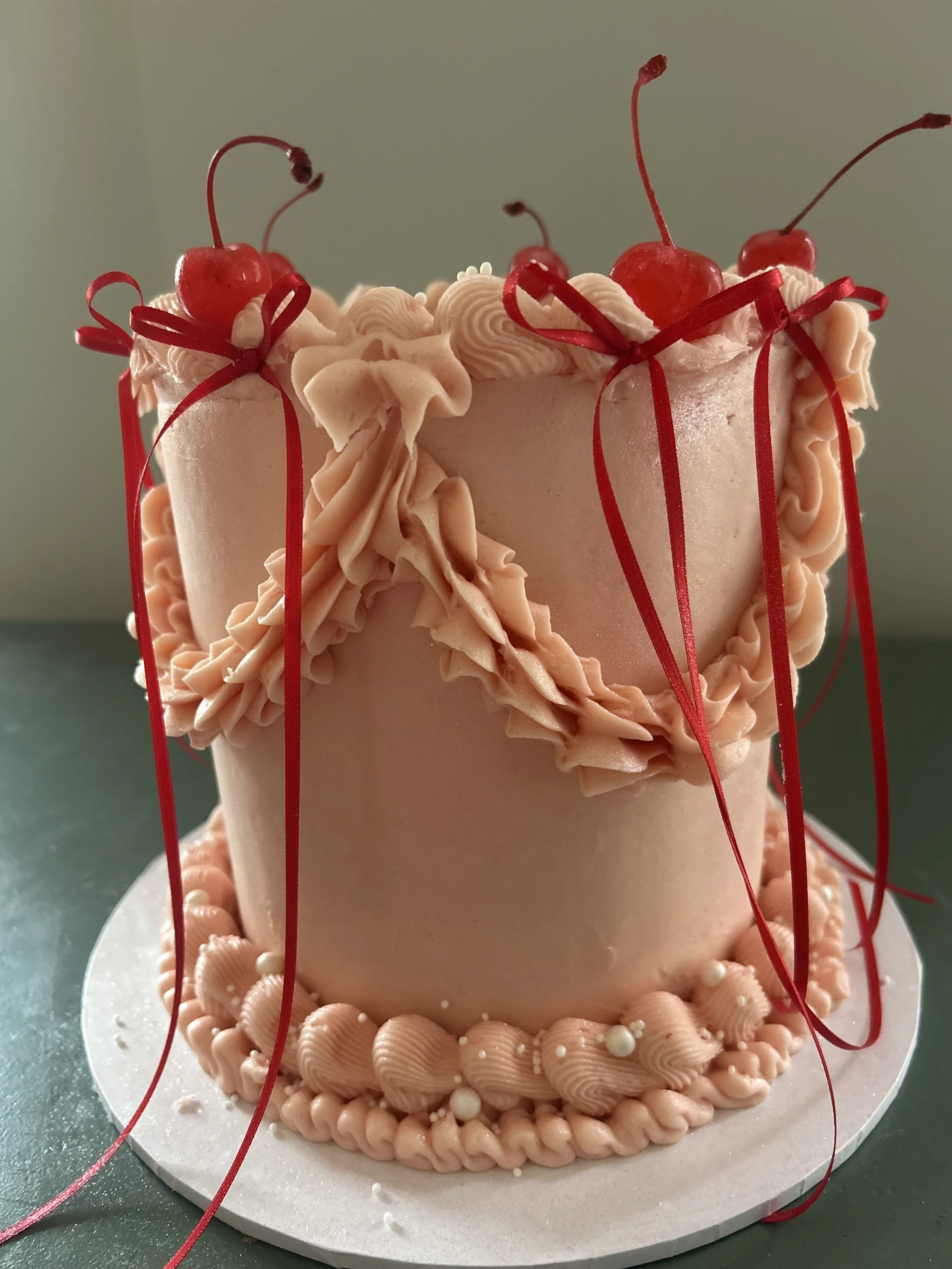 A pink birthday cake decorated with pink icing, pink pearls, and cherry toppings, with red ribbons cascading down the sides, placed on a white round cake board.