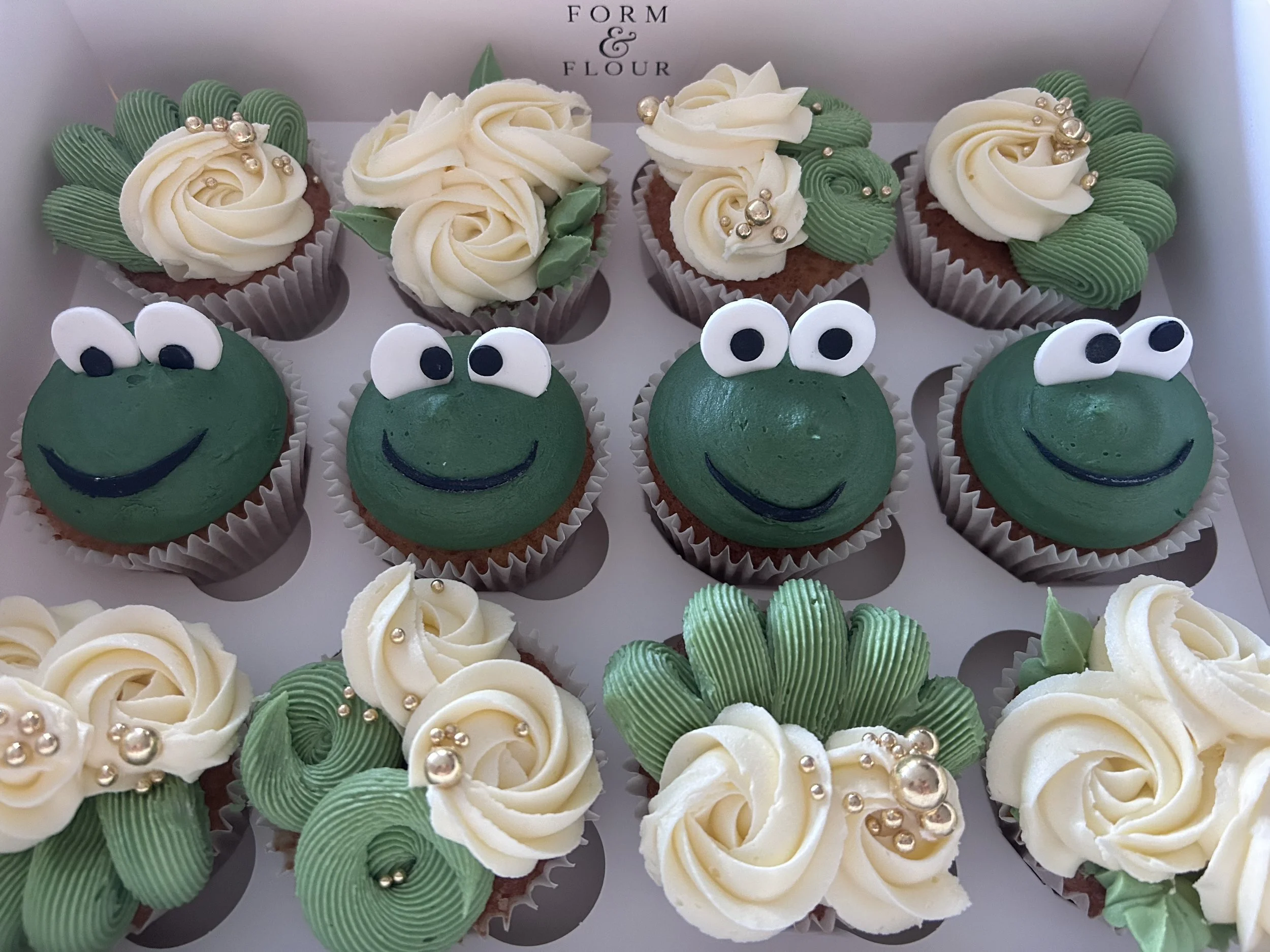 Cupcakes decorated with green frosting and white cream rosettes, some topped with silver edible pearls. Some cupcakes have frog faces with white eyes and black pupils, decorated with green frosting and black smiling mouths.