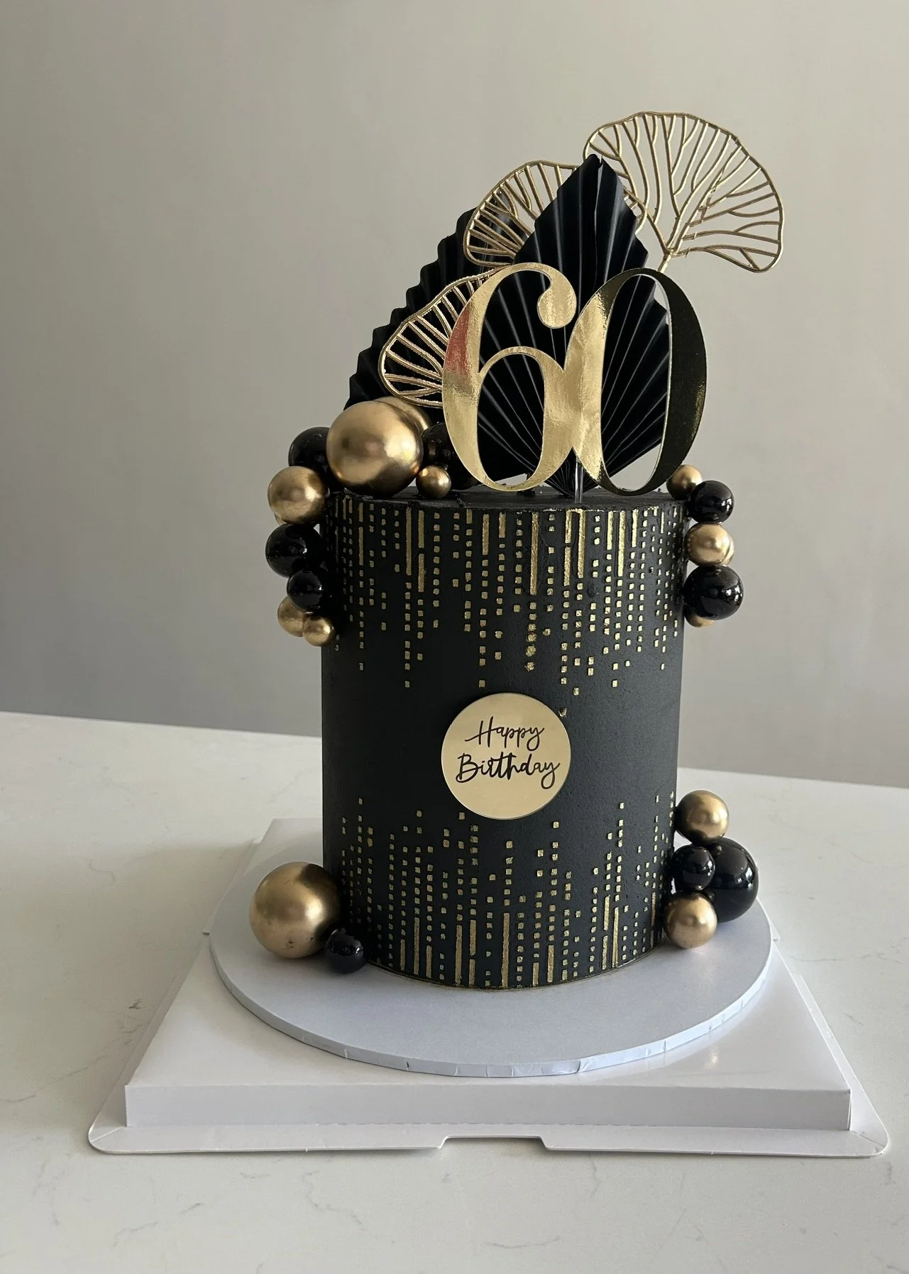 Black birthday cake with gold and black decorations, gold spheres, gold floral accents, and a large '60' topper. A gold 'Happy Birthday' message on the front of the cake.