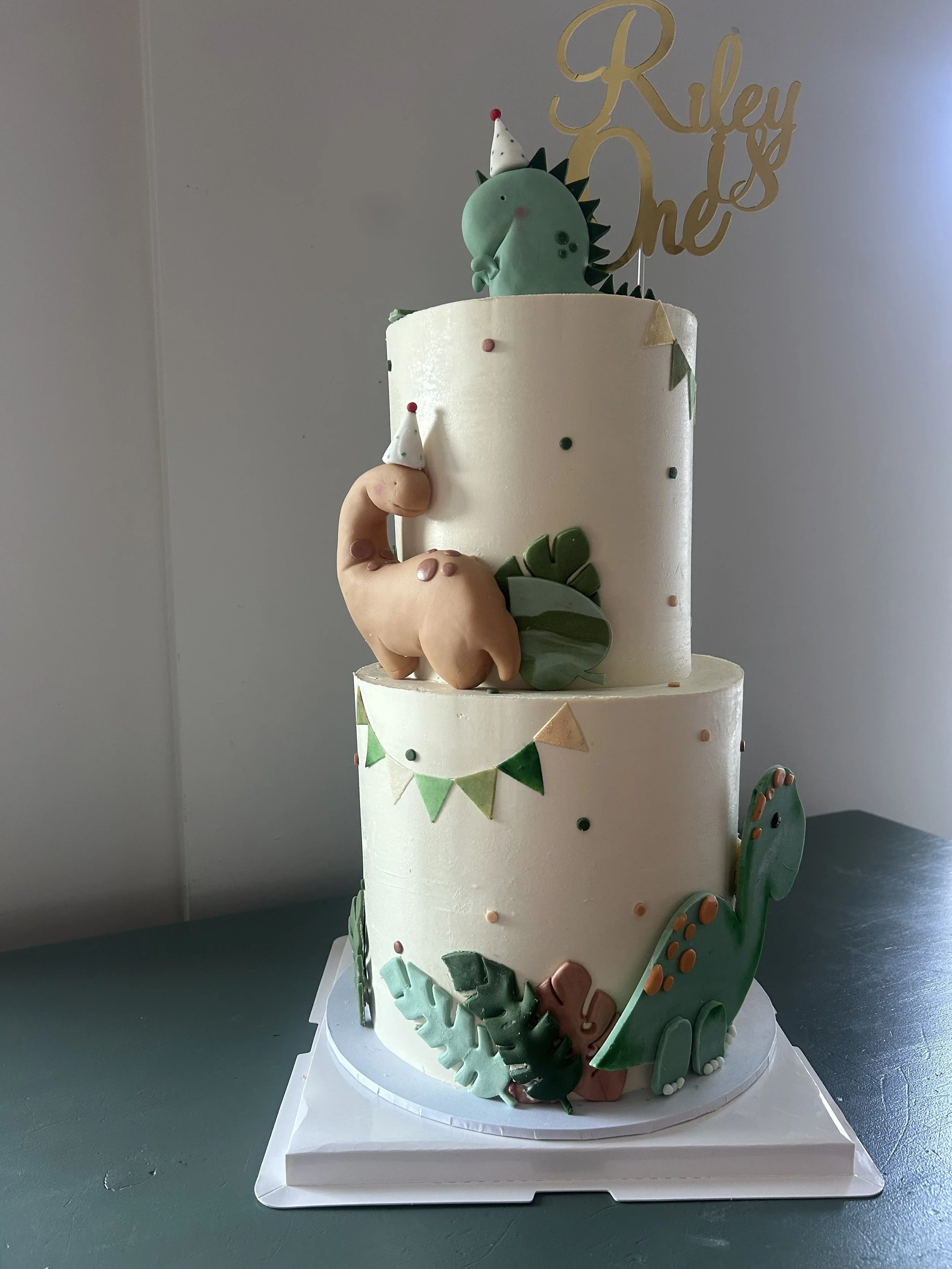 A multicolored birthday cake with a dinosaur theme. The cake has a dinosaur figurine wearing a party hat on top, a 'Riley & one' cake topper, and decorative elements like leaves and snowflakes around the sides.