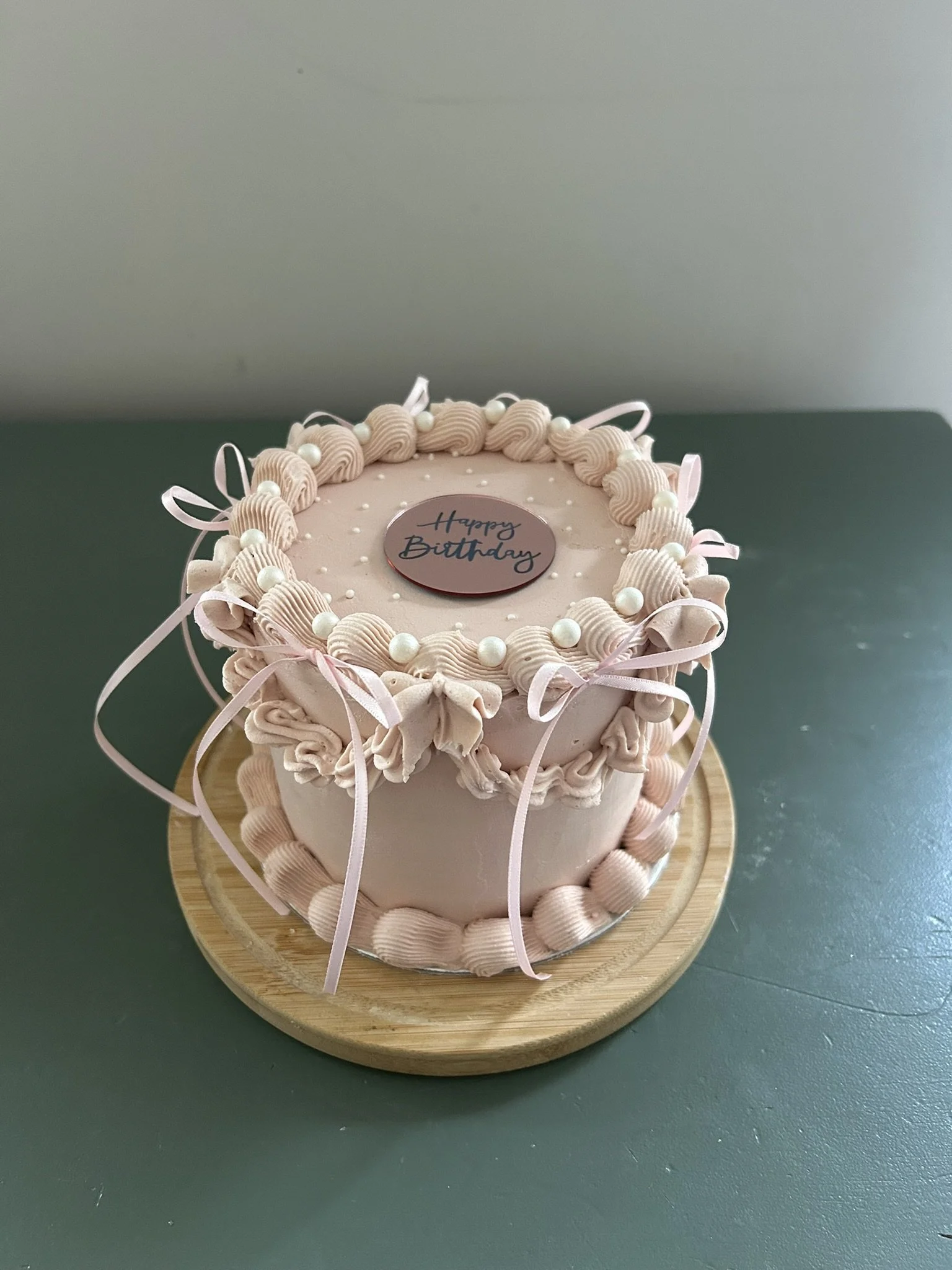 A pink birthday cake decorated with pink piped frosting, white beads, and ribbons, placed on a wooden cake stand with a "Happy Birthday" topper, against a gray background.