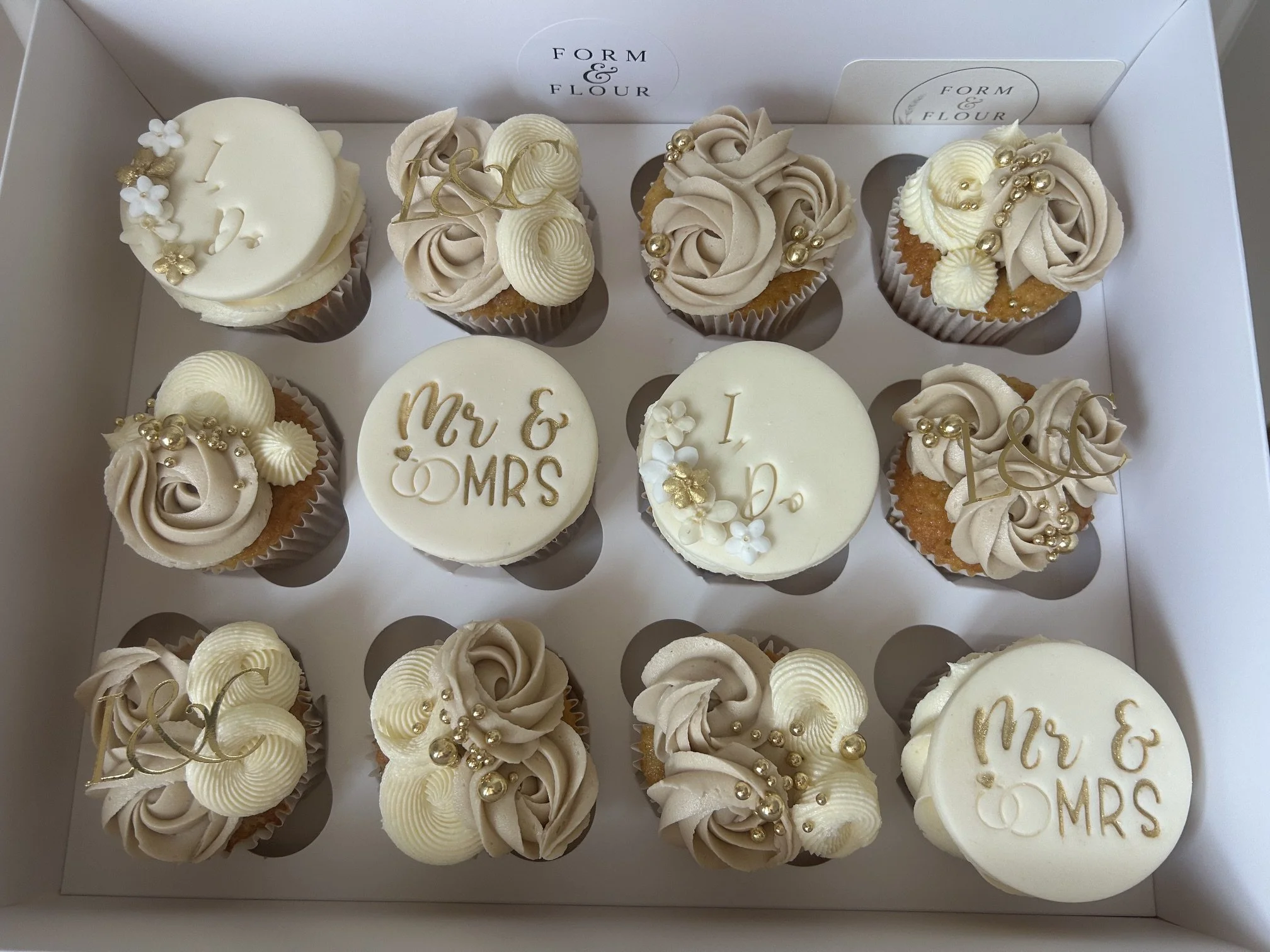 Box of decorated cupcakes with swirls of beige and white frosting, some topped with gold and white pearls, white flowers, and gold lettering that reads 'Mr & Mrs' and 'I Do.'