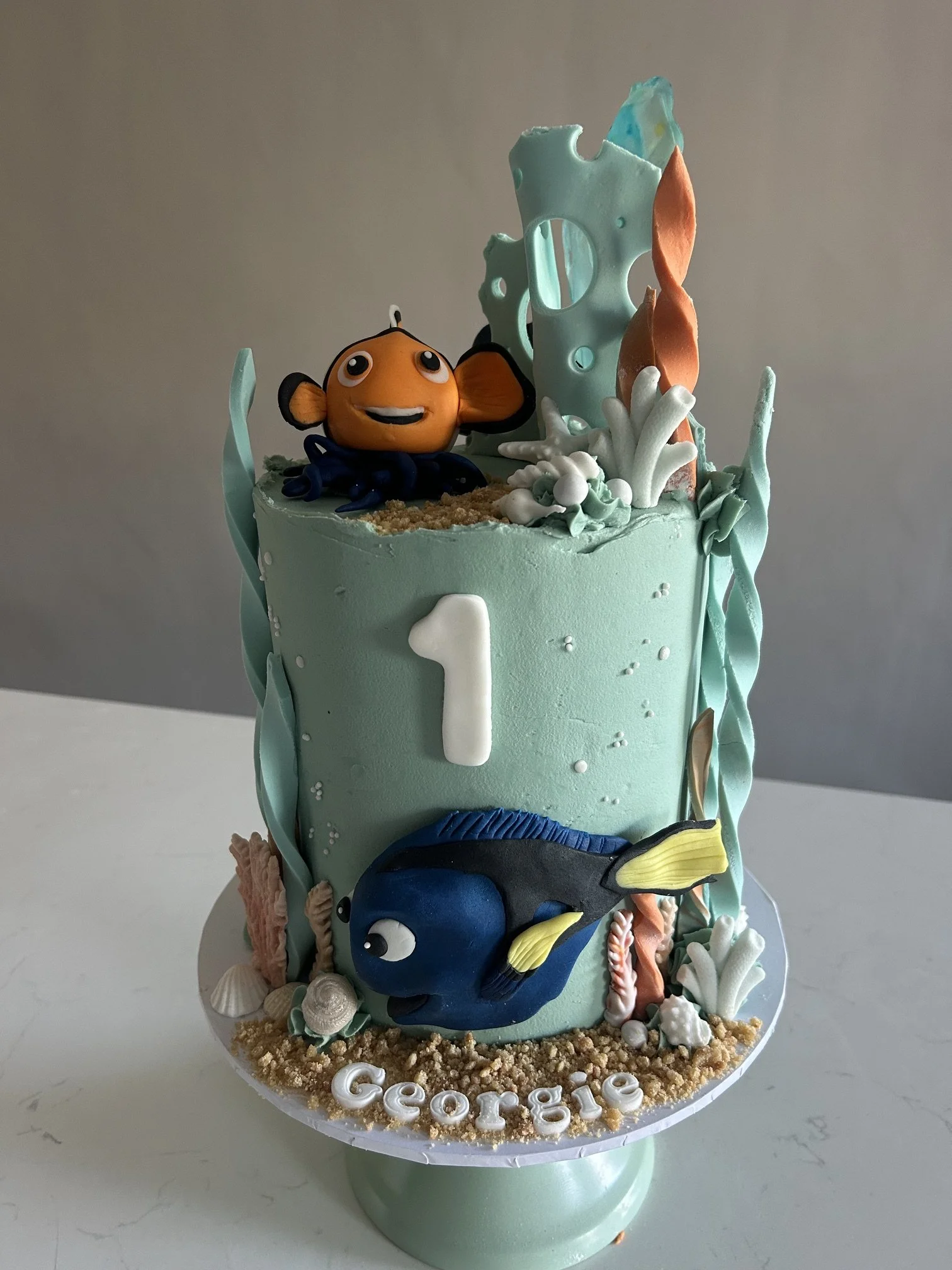 Decorated birthday cake with an underwater theme featuring fondant sea creatures including a clownfish and a blue tang, with shells and coral accents, and the name 'Georgie' written at the base.