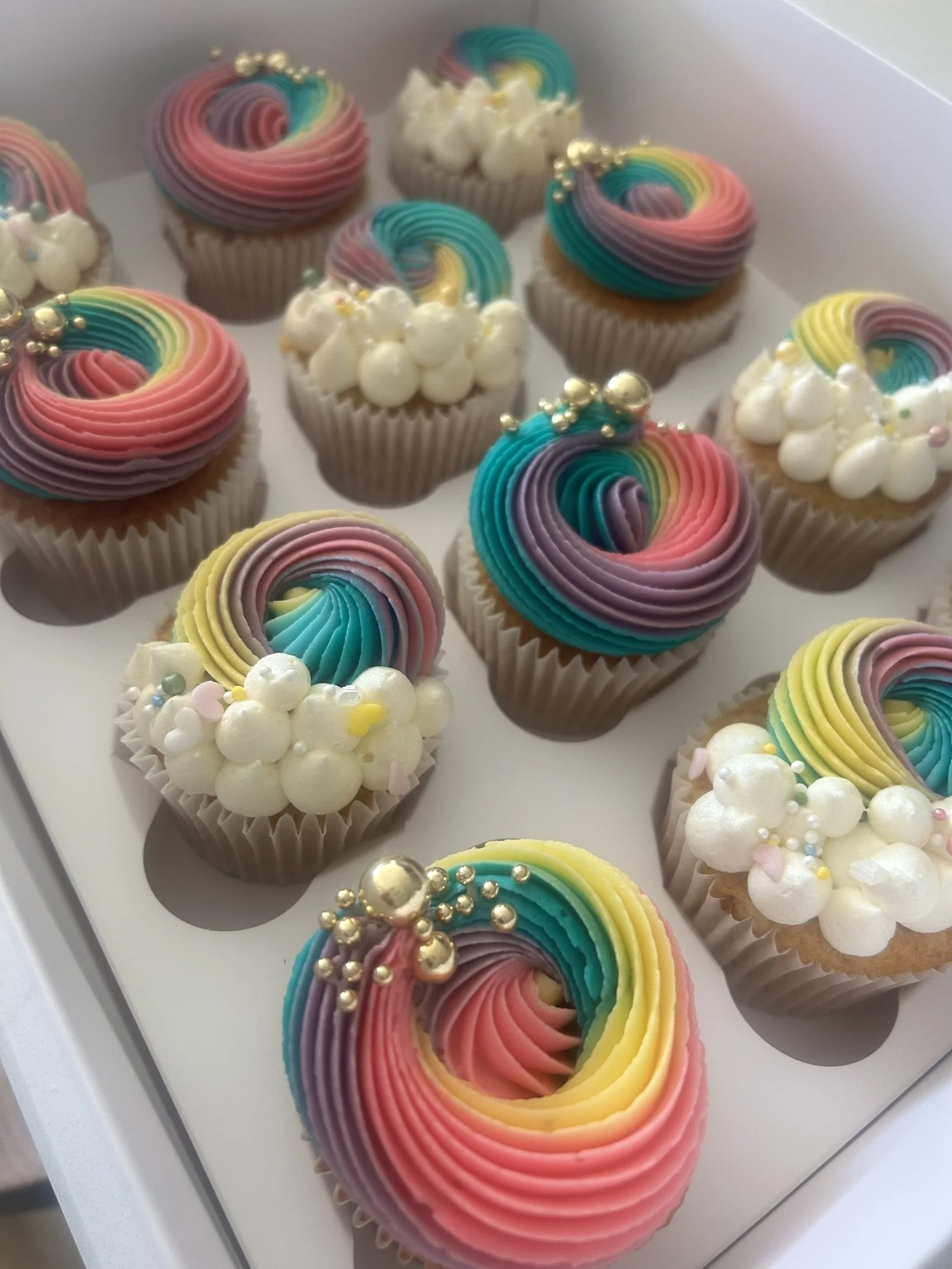 Colourful cupcakes decorated with rainbow  buttercream, white dollops of cream, and edible pearls on a white tray.