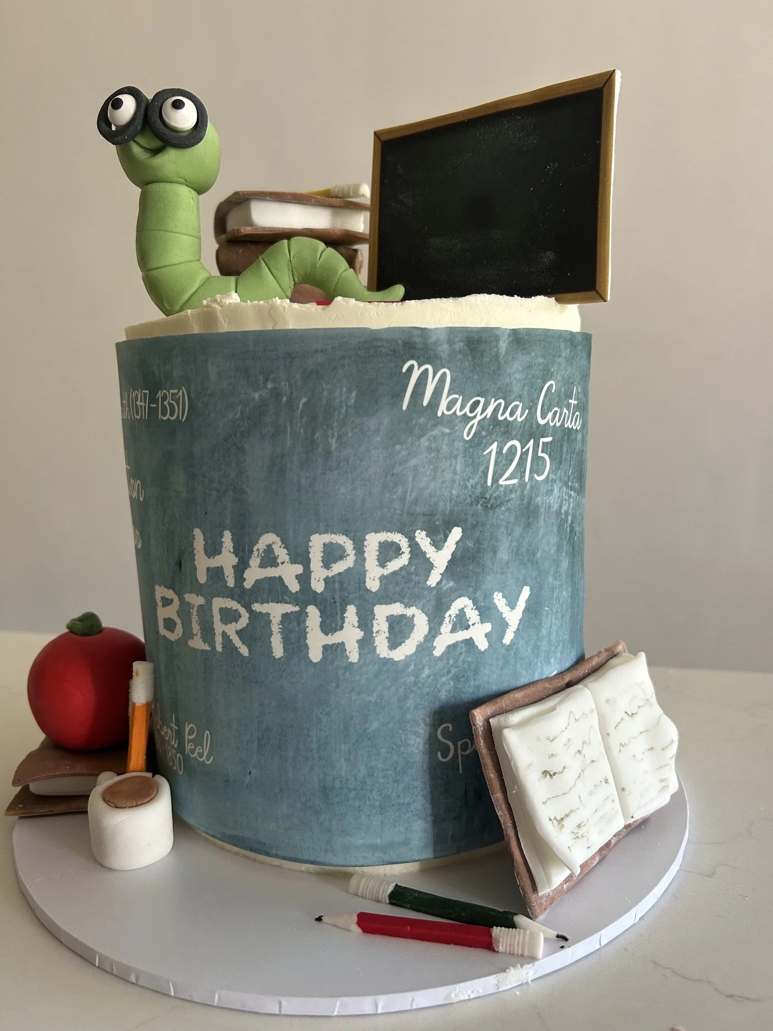 A birthday cake decorated with a green caterpillar figure with googly eyes, a chalkboard sign, and edible decorations like a strawberry, marshmallows, and cookies, with birthday message text on the cake.