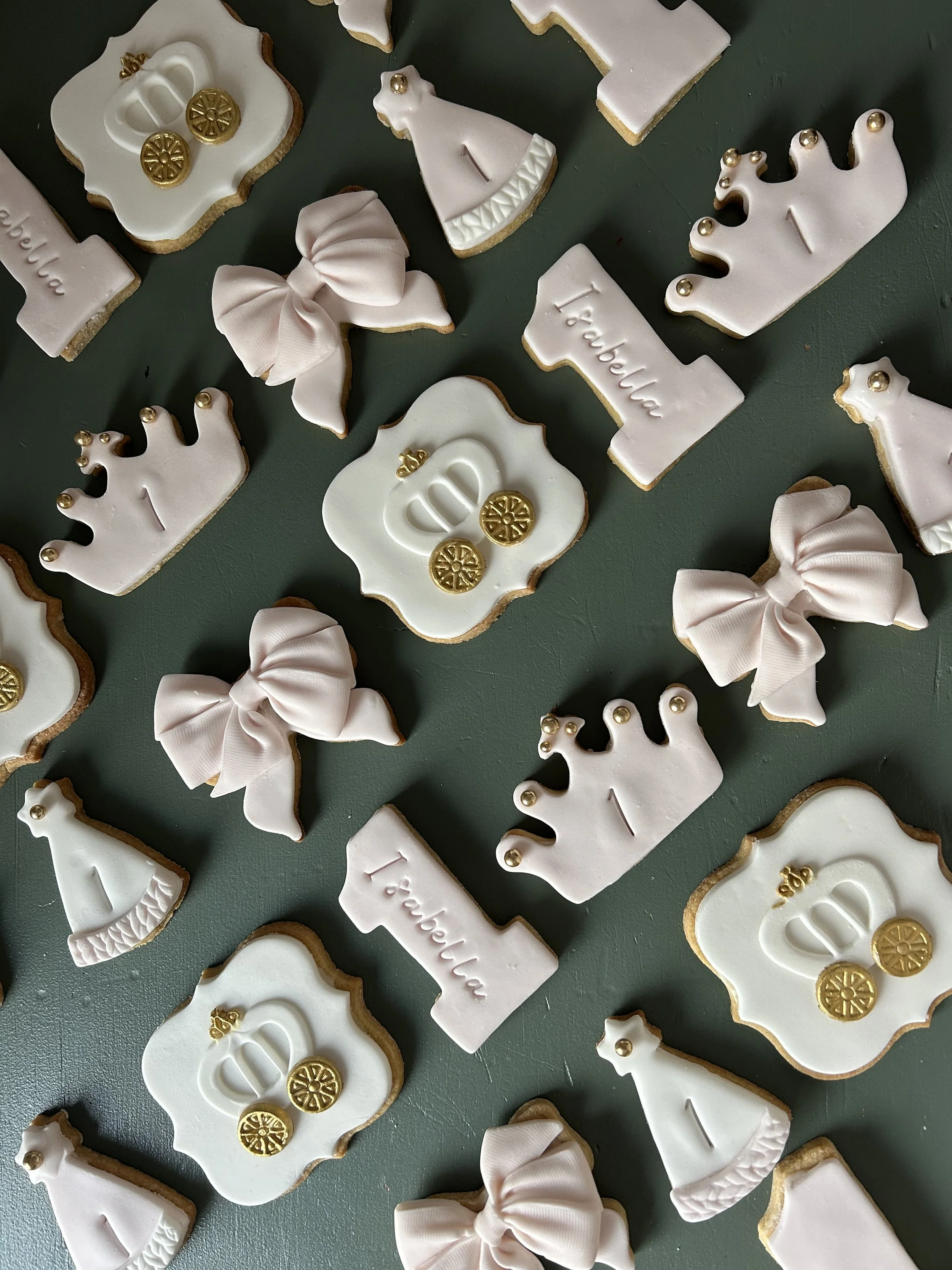 Decorated sugar cookies with white icing, gold accents, and pink bows, arranged on a dark surface.