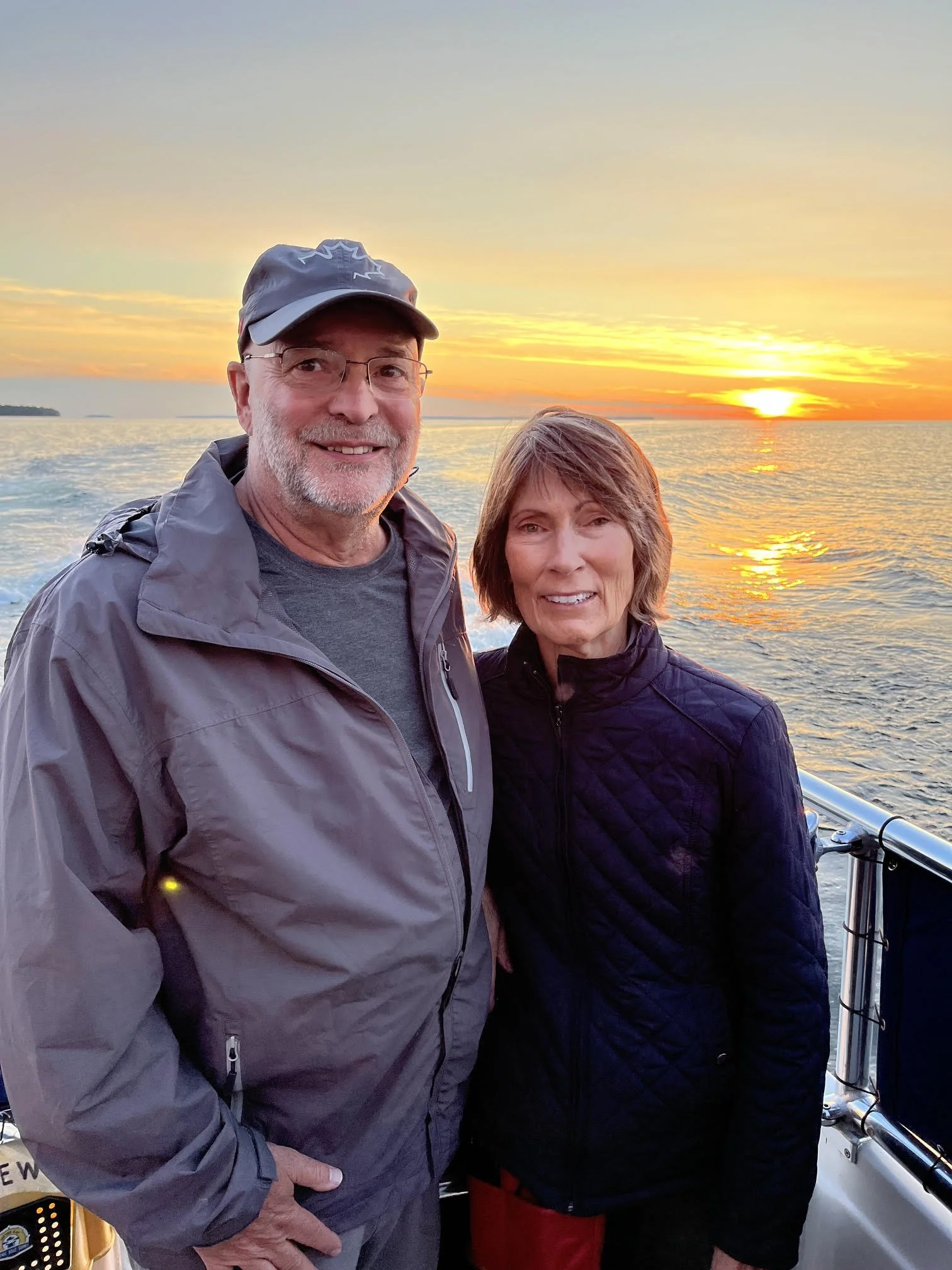 A smiling middle-aged man in a gray jacket and cap and a woman in a black quilted jacket and red shorts take a selfie on a boat at sunset, with the ocean and a colorful sky in the background.