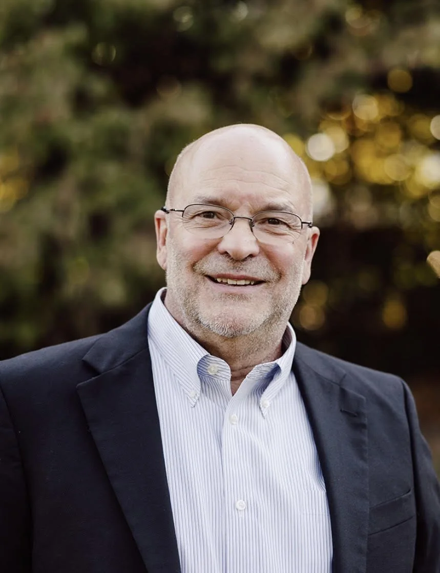 A middle-aged man with glasses and a trimmed beard, wearing a navy blazer and a light blue collared shirt, smiling outdoors with blurred trees and golden sunlight in the background.