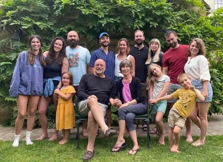 Family group photo outdoors in front of green foliage, with adults and children smiling and posing.