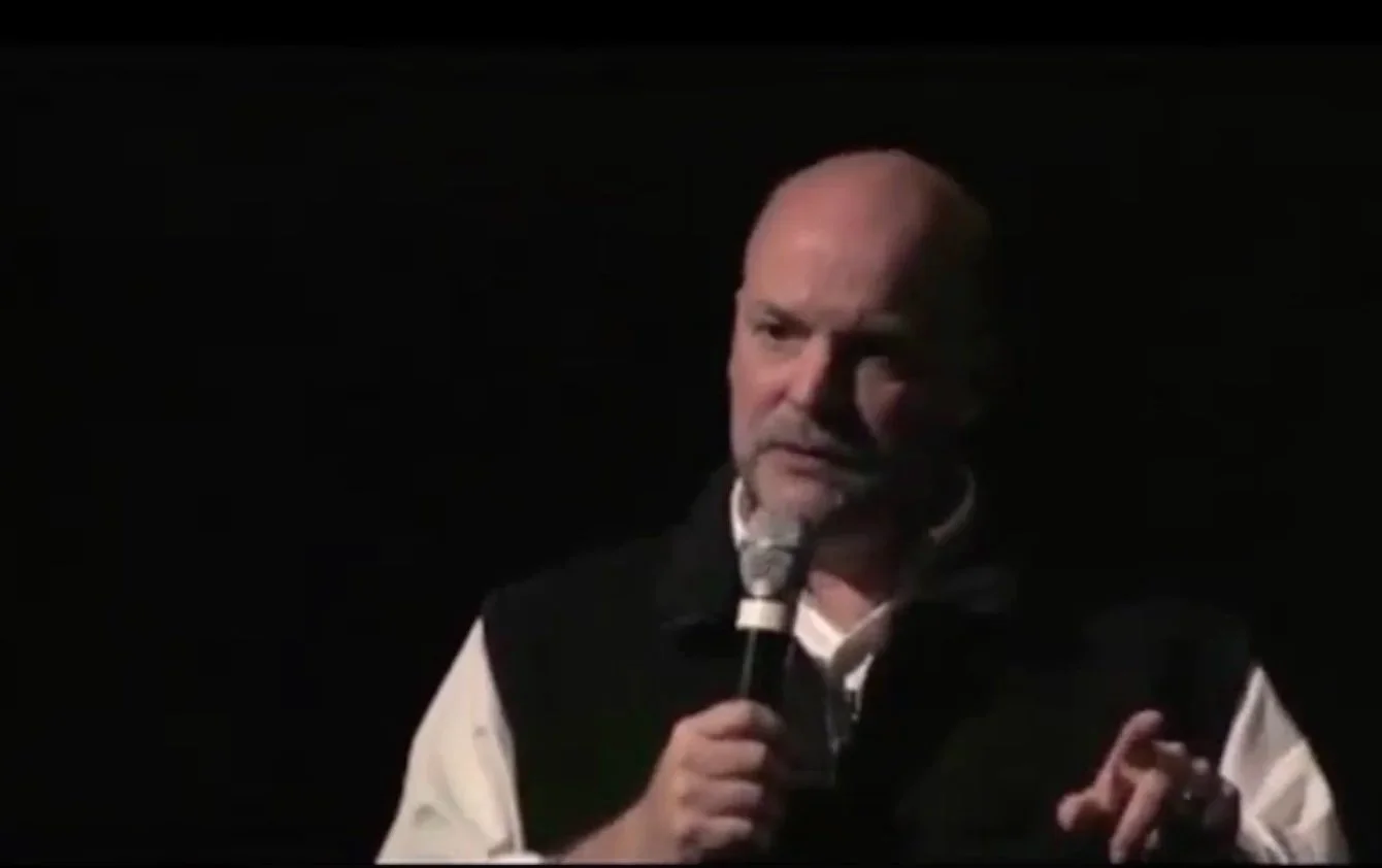 Bald man holding a microphone, speaking on stage in a dark setting.