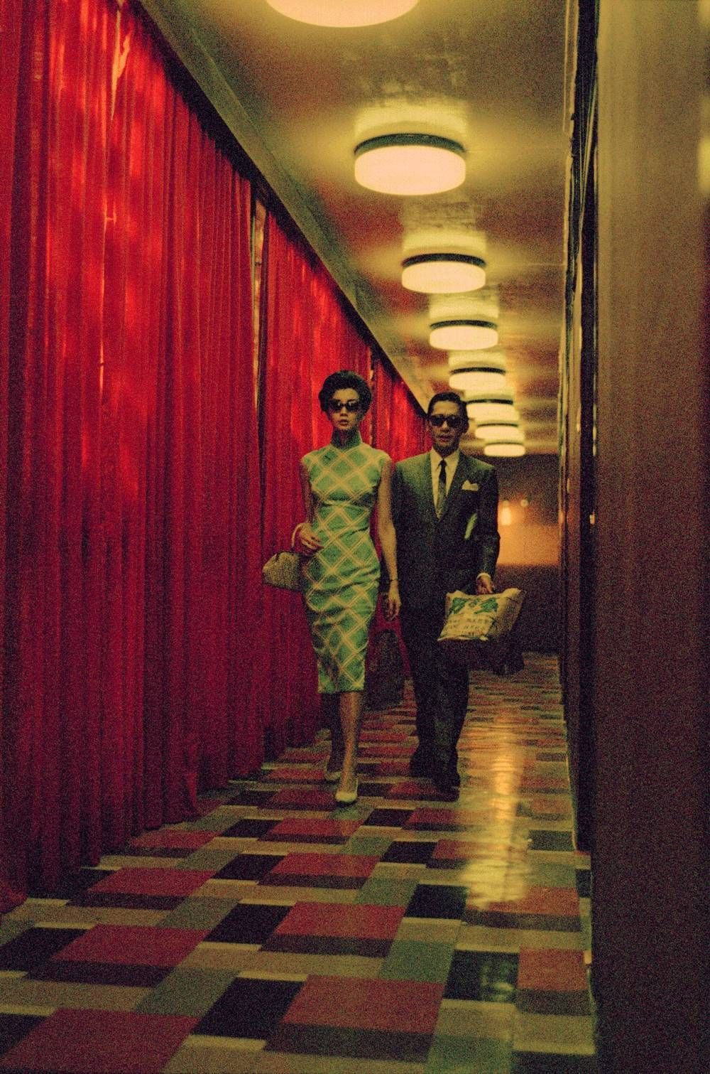 A man and woman in vintage 1960s attire walking down a hallway illuminated by overhead lights, with red curtains on one side and a checkered floor.