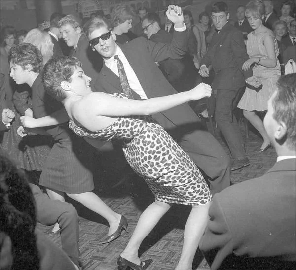 Black and white photo of a dance party with young people dancing closely together, some holding each other, in a retro style setting.