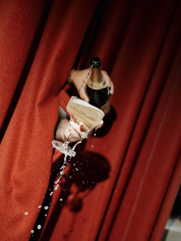 A person holding a champagne bottle with a chain around the neck, a glass with champagne, and champagne spilling out, against a red curtain background.