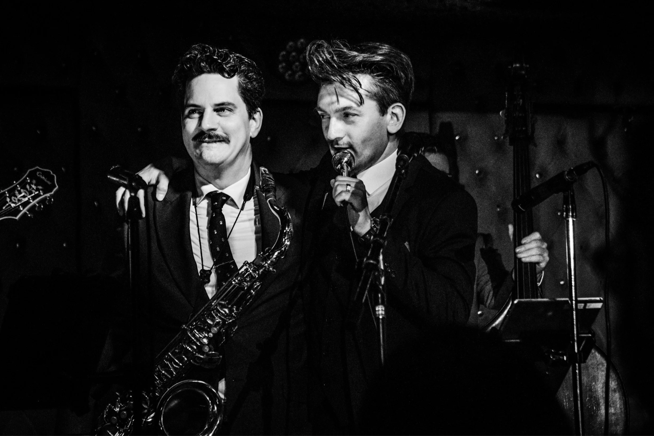 Two men performing on stage in a black-and-white photo. One holds a saxophone, and the other is singing into a microphone. The man on the left has dark, wavy hair, a mustache, and is dressed in a suit and tie. The man on the right has styled hair, a white shirt, black jacket, and appears to be singing.