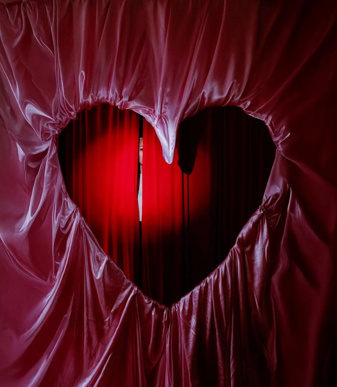 A person peeking through a heart-shaped opening in red curtains on a stage.