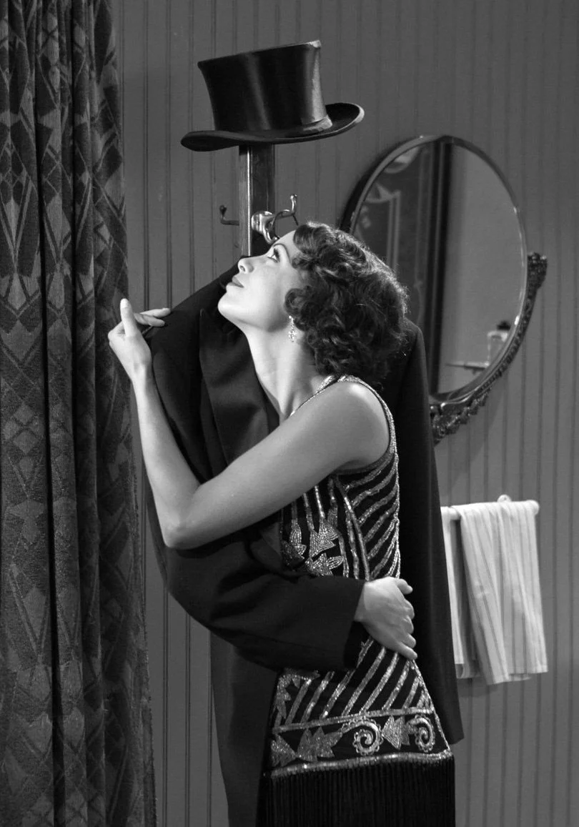 A vintage black and white photo of a woman with curly hair, holding a coat in a room with wooden paneling, a round mirror, and a towel hanging on a rack.