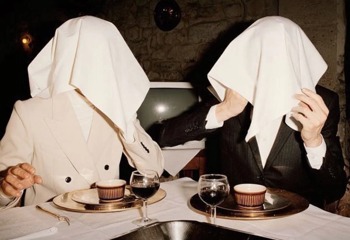 Two people dining at a table with white tablecloth, glassware, and bowls, both with cloths over their heads, dressed in formal attire, in a dimly lit restaurant with brick walls.