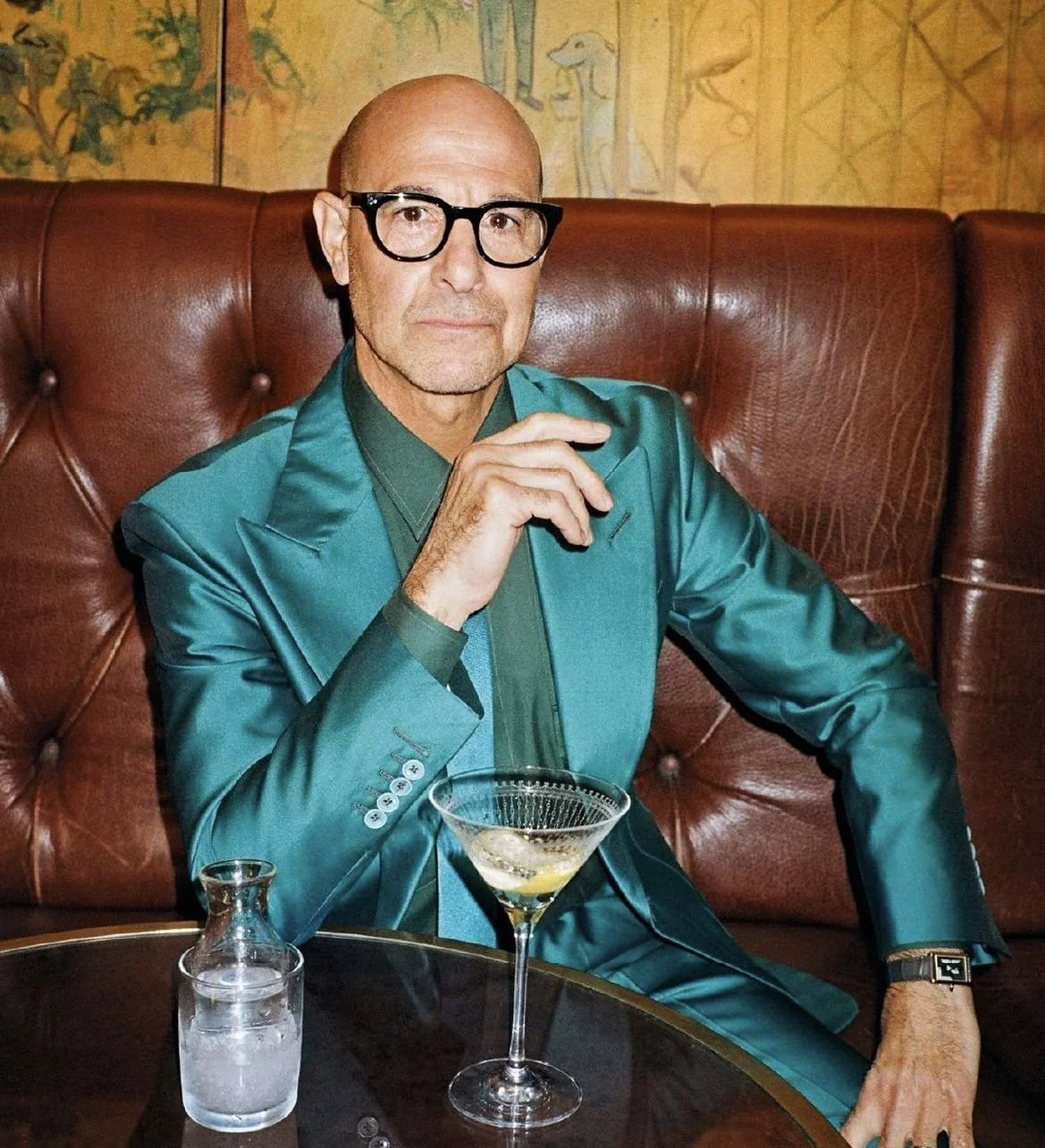 A man sitting in a booth at a restaurant or bar, wearing glasses and a teal suit, holding a cocktail glass, with a pitcher of water on the table.
