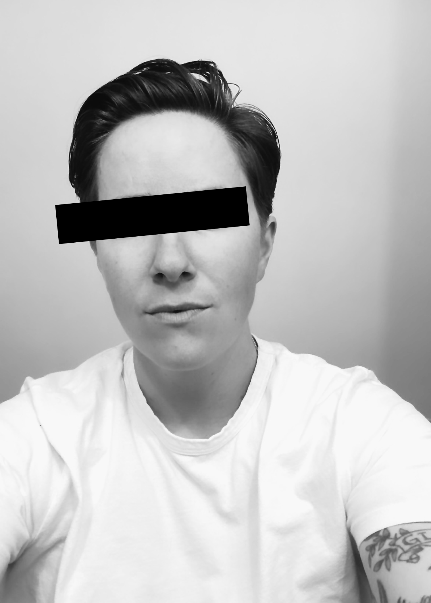 Black and white photo of a person with a side part hairstyle, wearing a plain white T-shirt, showing a tattoo on their right arm with some floral design.