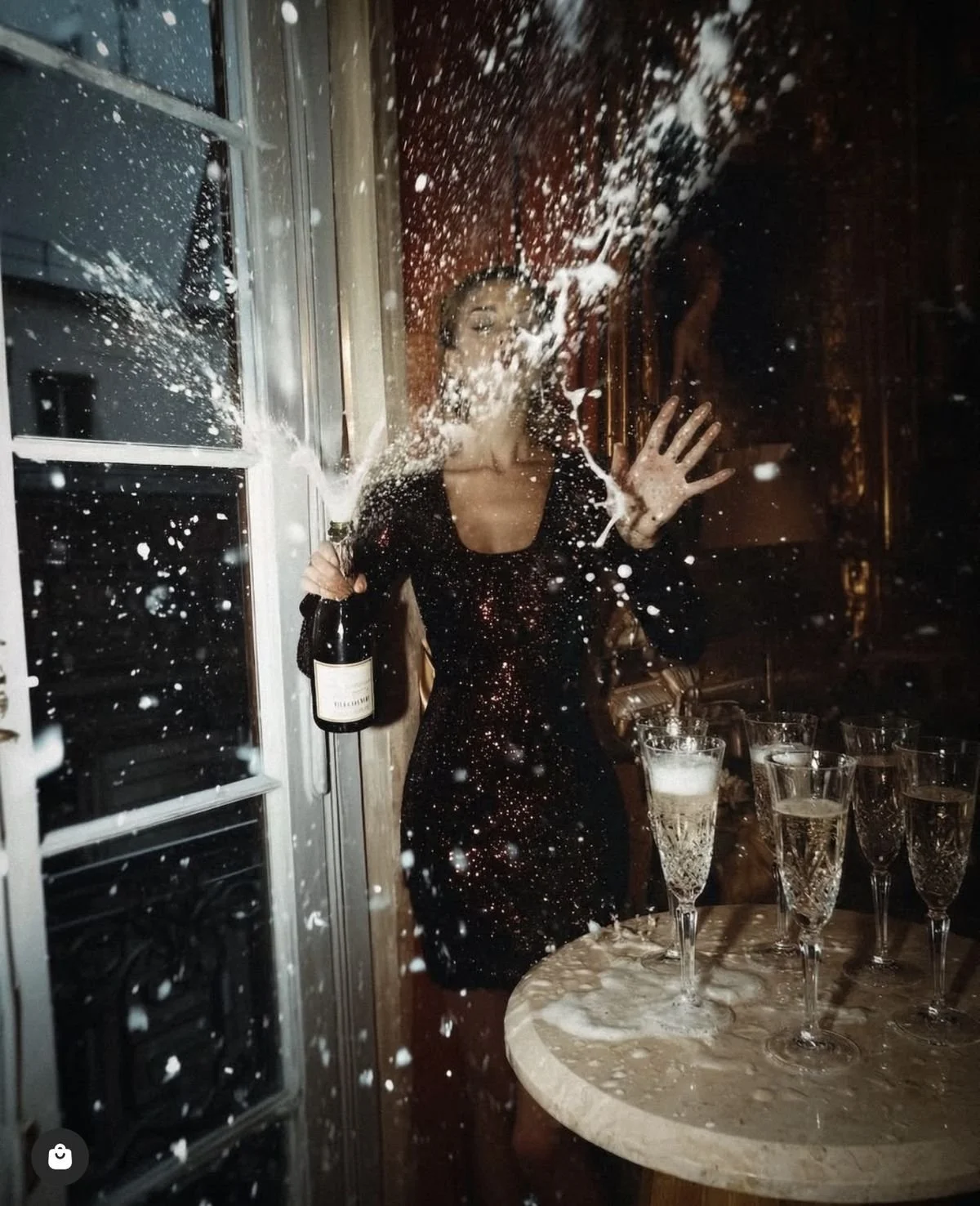 Woman in a black dress celebrating with a champagne bottle, splashing foam in a window at night, with several glasses of champagne on a nearby table.