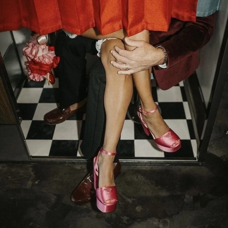A man and woman pictured in an elevator, with the woman sitting on the man's lap. The woman is wearing pink high heels, and the man is wearing brown dress shoes. The woman’s gown is lifted slightly, revealing her legs. The background shows black and white checkered flooring.