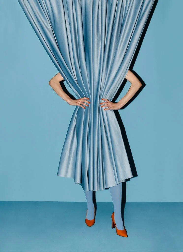 Person hiding behind a blue curtain with only legs visible, wearing orange high heels and striped tights, posing with hands on hips against a plain blue background.