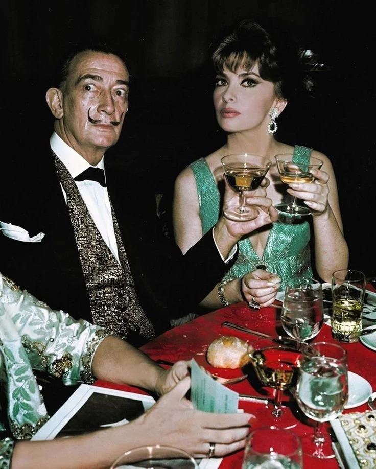 A man and a woman dressed in formal attire at a dinner party, holding glasses of drinks, with a table set with glassware and food.