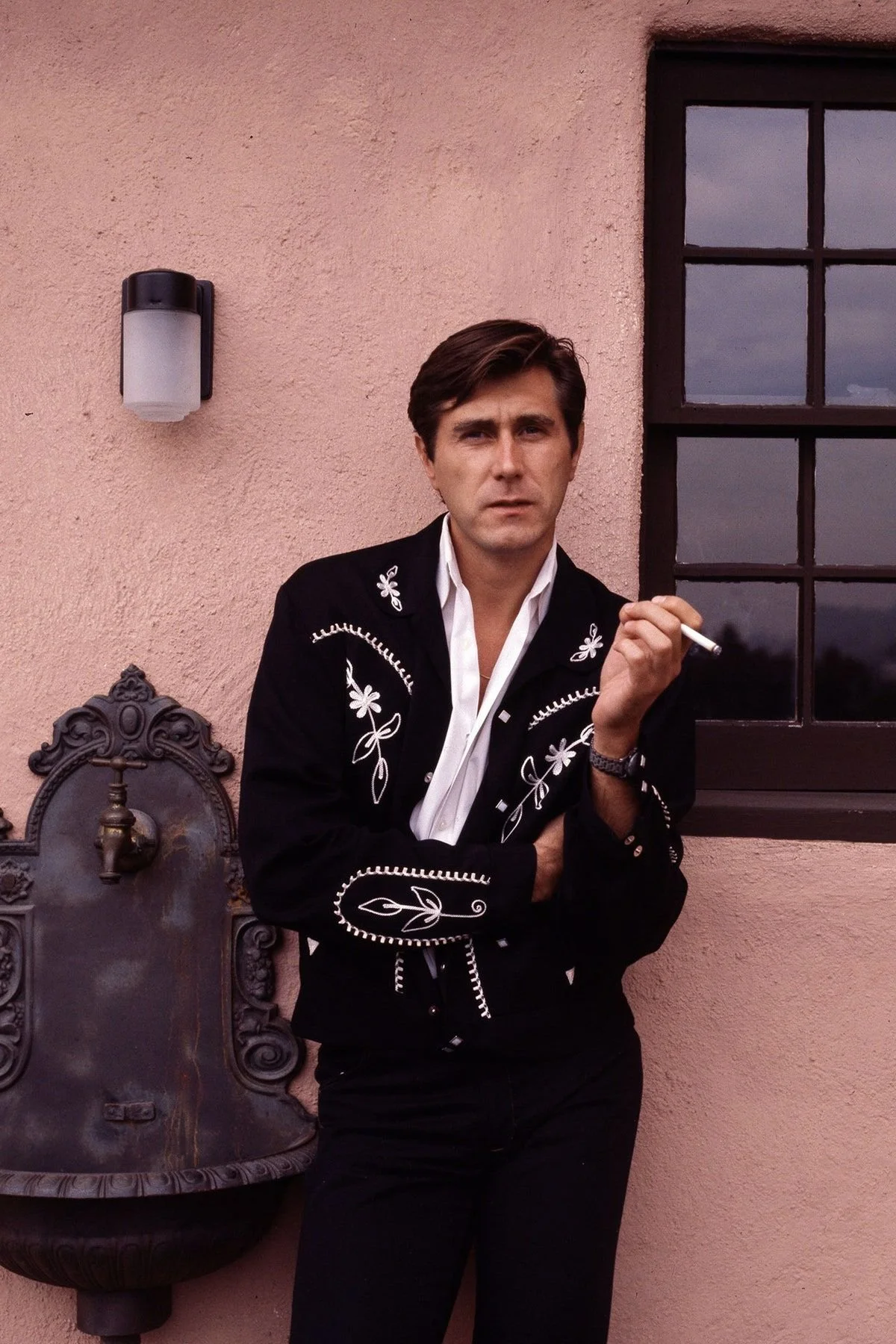 A man with dark hair, wearing a black embroidered jacket, white shirt, and black pants, holding a cigarette, stands in front of a pink wall with a window and an antique water fountain.