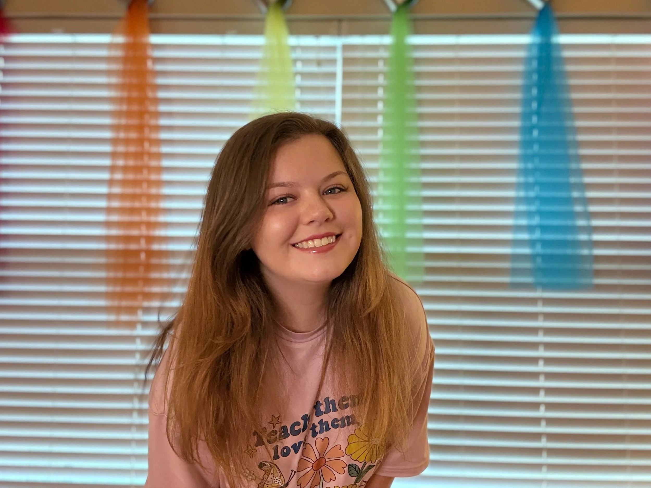 Young woman with long, reddish-brown hair smiling in front of a colorful striped backdrop of vertical blinds painted in orange, yellow, green, and blue.