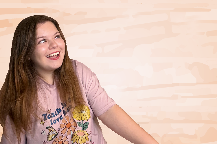 A smiling young woman with long brown hair, wearing a pink T-shirt with floral and butterfly graphics, looking up and to her right against a light beige brick wall background.
