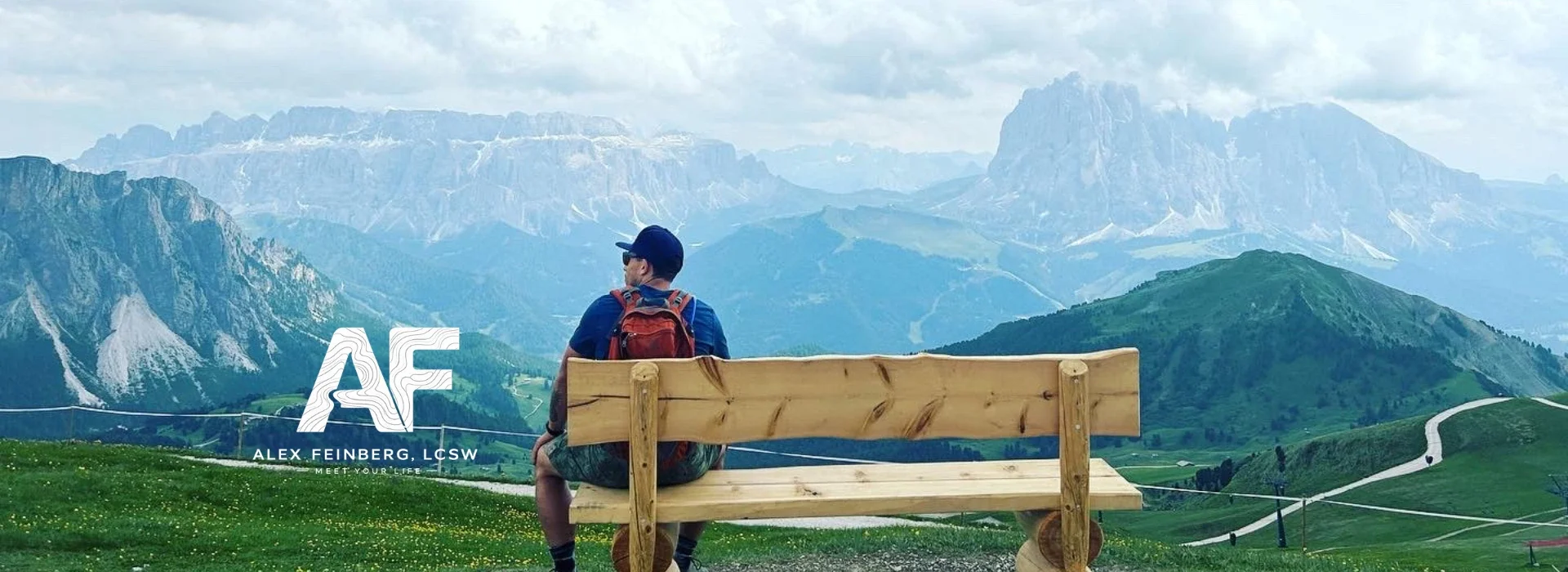 A person sitting on a wooden bench overlooking green rolling hills and mountains under a cloudy sky.