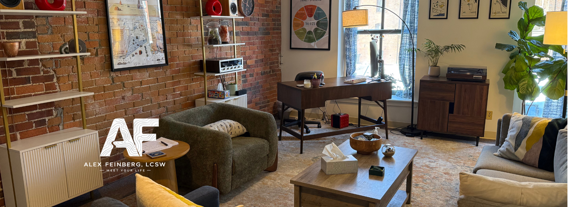 A cozy living room with a brick accent wall, a desk with a computer, a comfortable green armchair, a sofa with pillows, and various decorative items and plants.