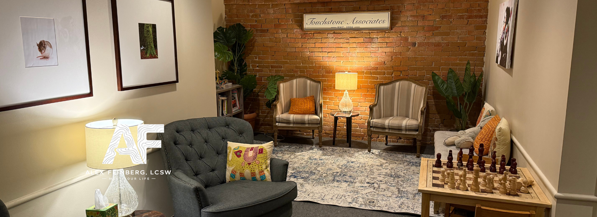 Cozy waiting room with gray tufted armchair, two striped armchairs, a sofa with cushions, a wooden chess table with chess pieces, a lamp, indoor plants, framed pictures on the wall, and a sign reading "Touchstone Associates" on a brick wall.