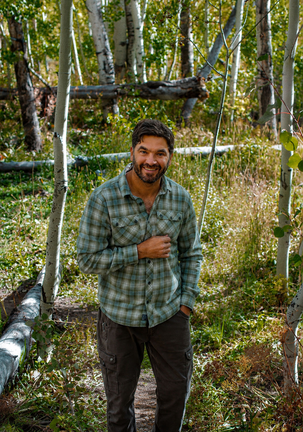 A man with dark hair and a beard smiling in a forest, wearing a plaid shirt and dark pants.