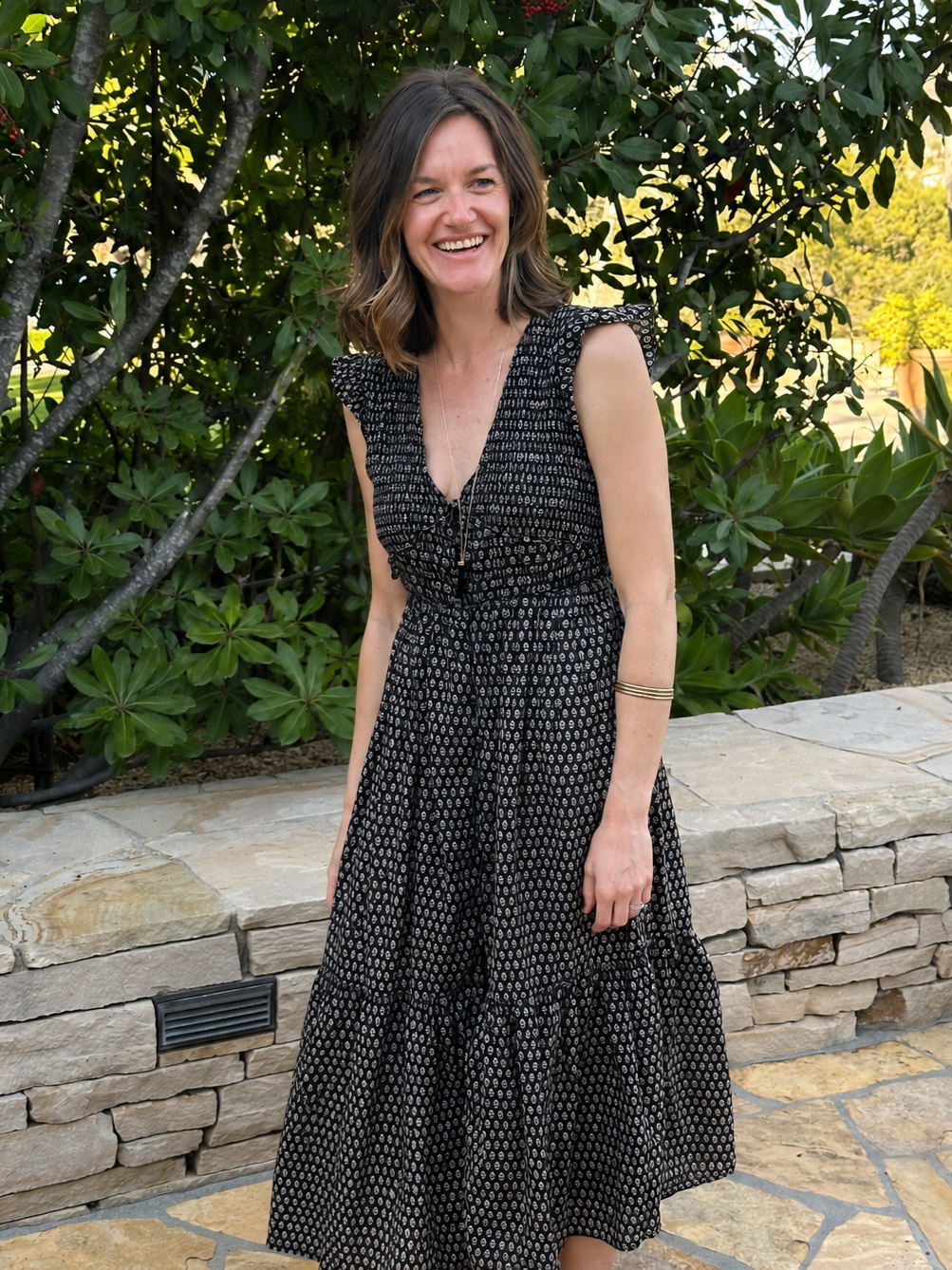 A smiling woman with shoulder-length brown hair in a black patterned dress standing outdoors in front of green leafy bushes and trees.