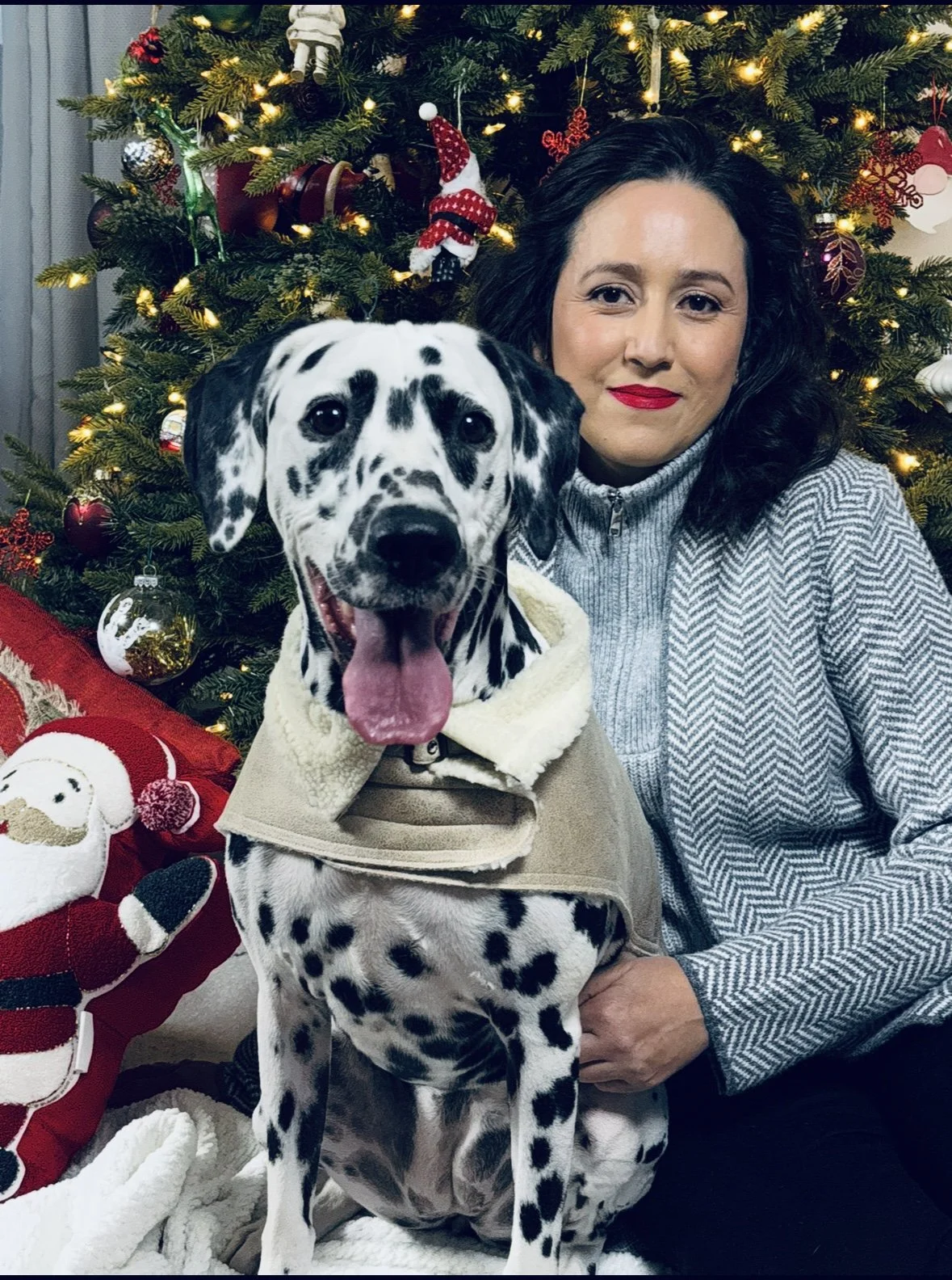 A woman with black hair and red lipstick smiling while sitting next to a Dalmatian puppy in front of a decorated Christmas tree with lights and ornaments.