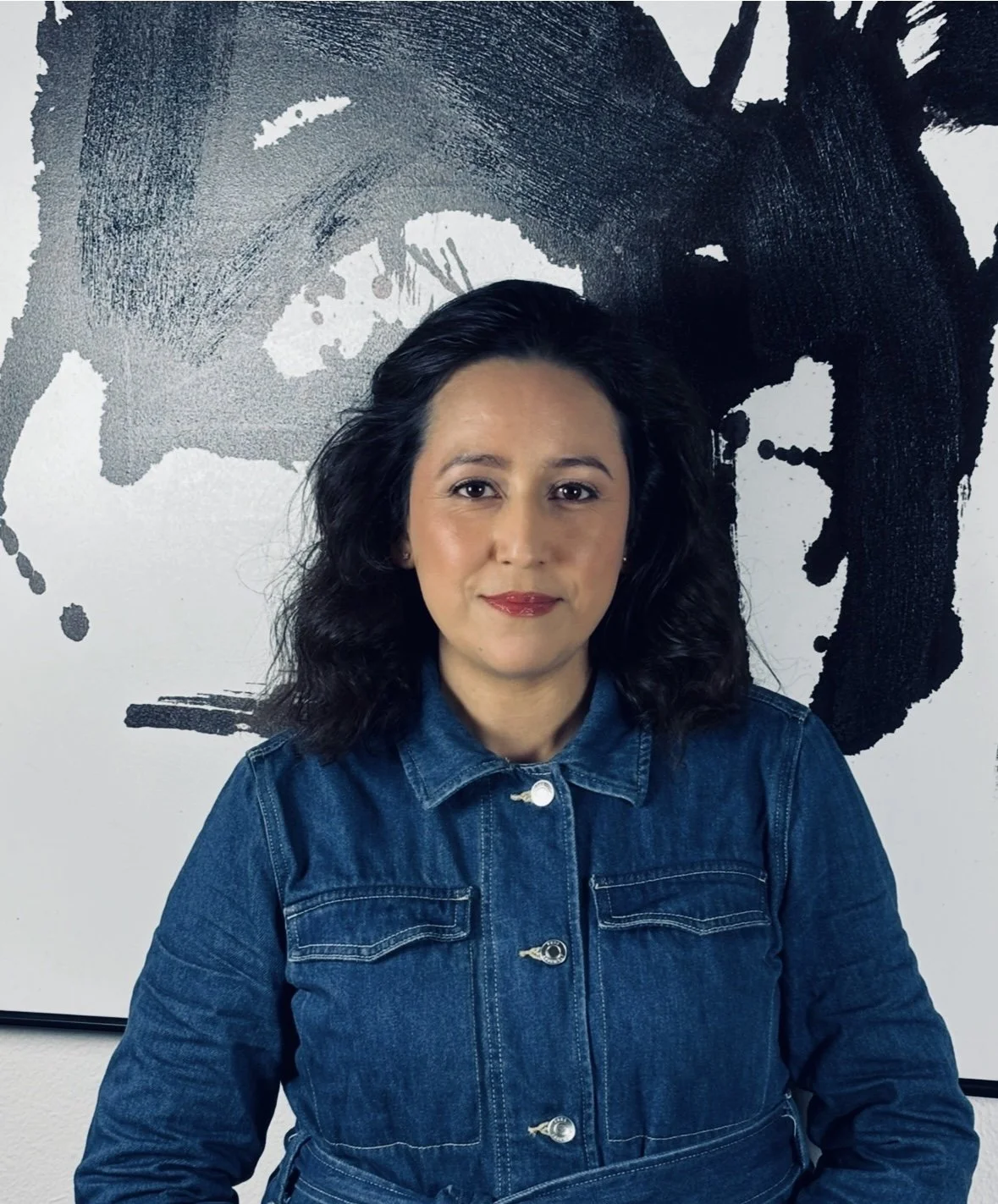 A woman with curly black hair wearing a denim jacket, standing in front of a black and white abstract artwork.