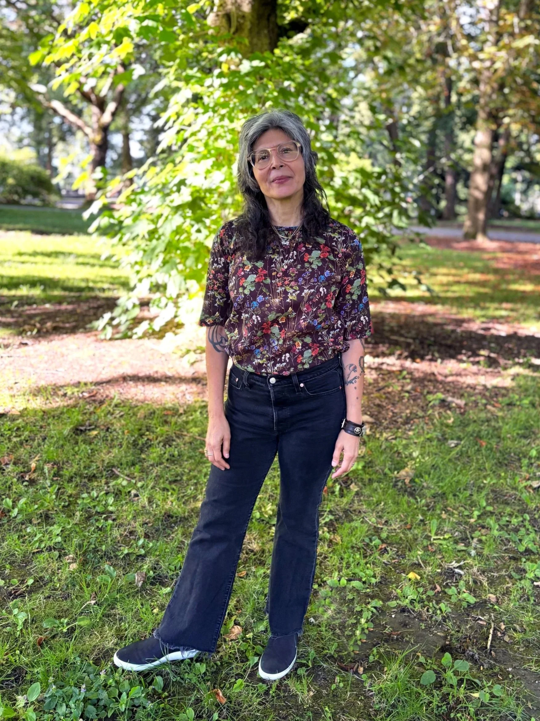 A woman with wavy gray and black hair, wearing glasses with clear frames, a shirt with a colorful floral pattern. Judy Almazan Stuhmer uses EMDR, IFS informed. Helps with complex PTSD, anxiety, relationships, toxic work places, identity and belonging