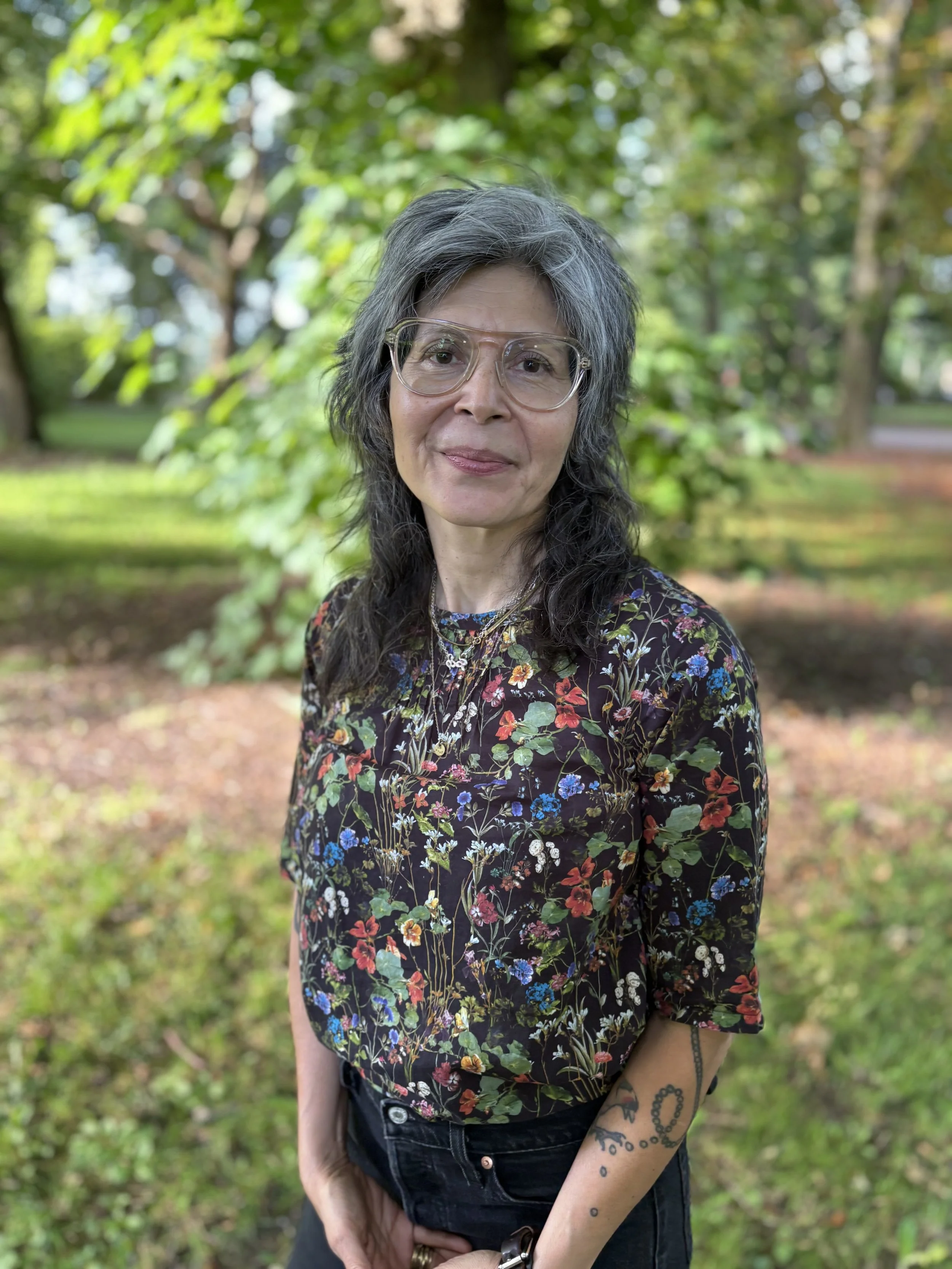 A woman with long dark hair and glasses standing outdoors in a park with green trees and grass in the background. Judy Almazan Stuhmer uses EMDR, IFS informed. Helps with complex PTSD, anxiety, relationships, toxic work places, identity and belonging
