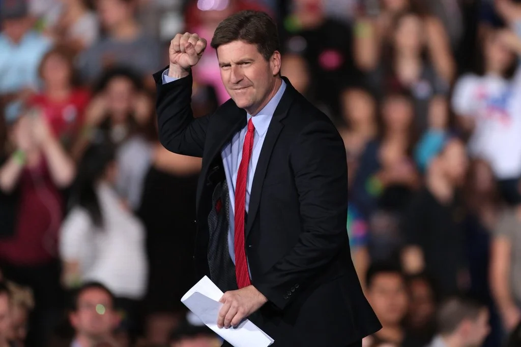 A man in a suit making a fist gesture with a crowd in the background.