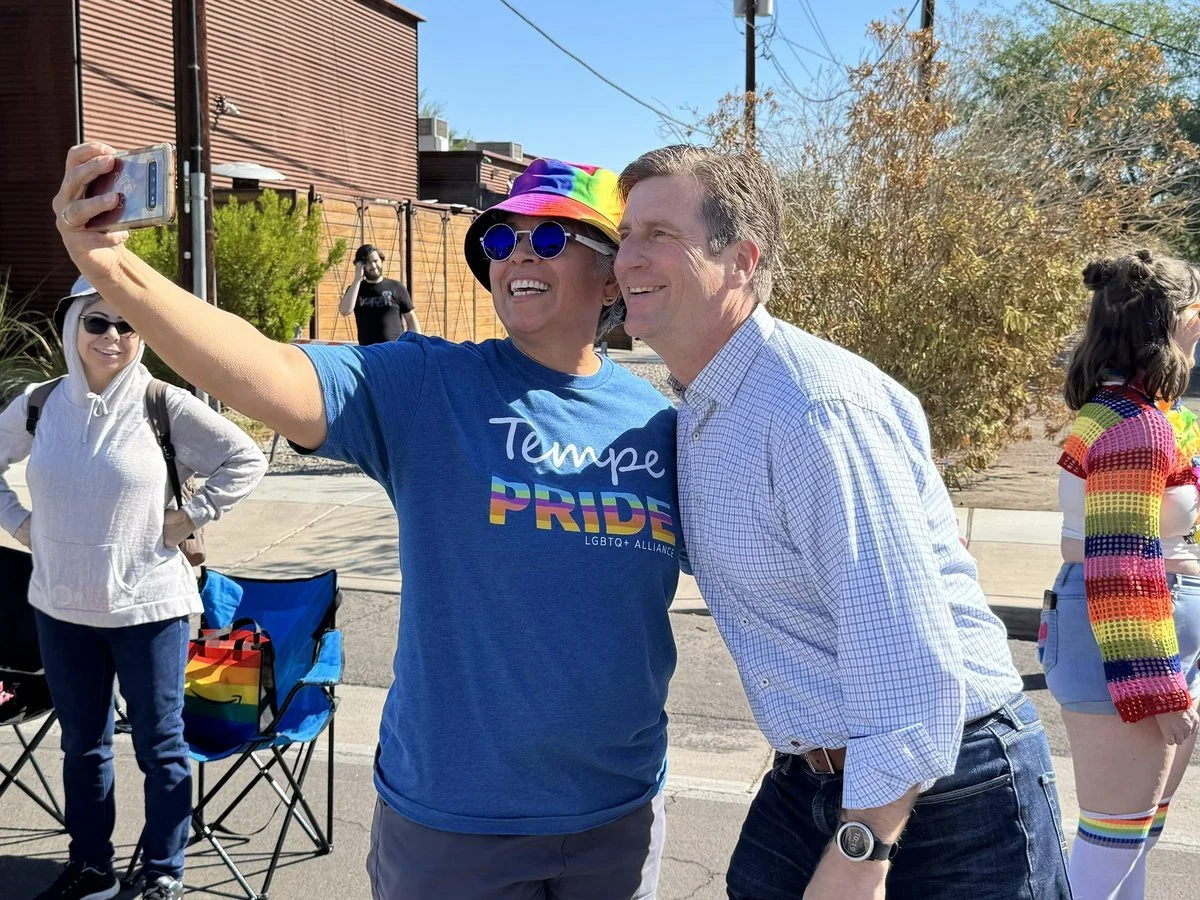 Two people taking a selfie at a pride event, with rainbow-themed accessories and clothing, and others gathered around.
