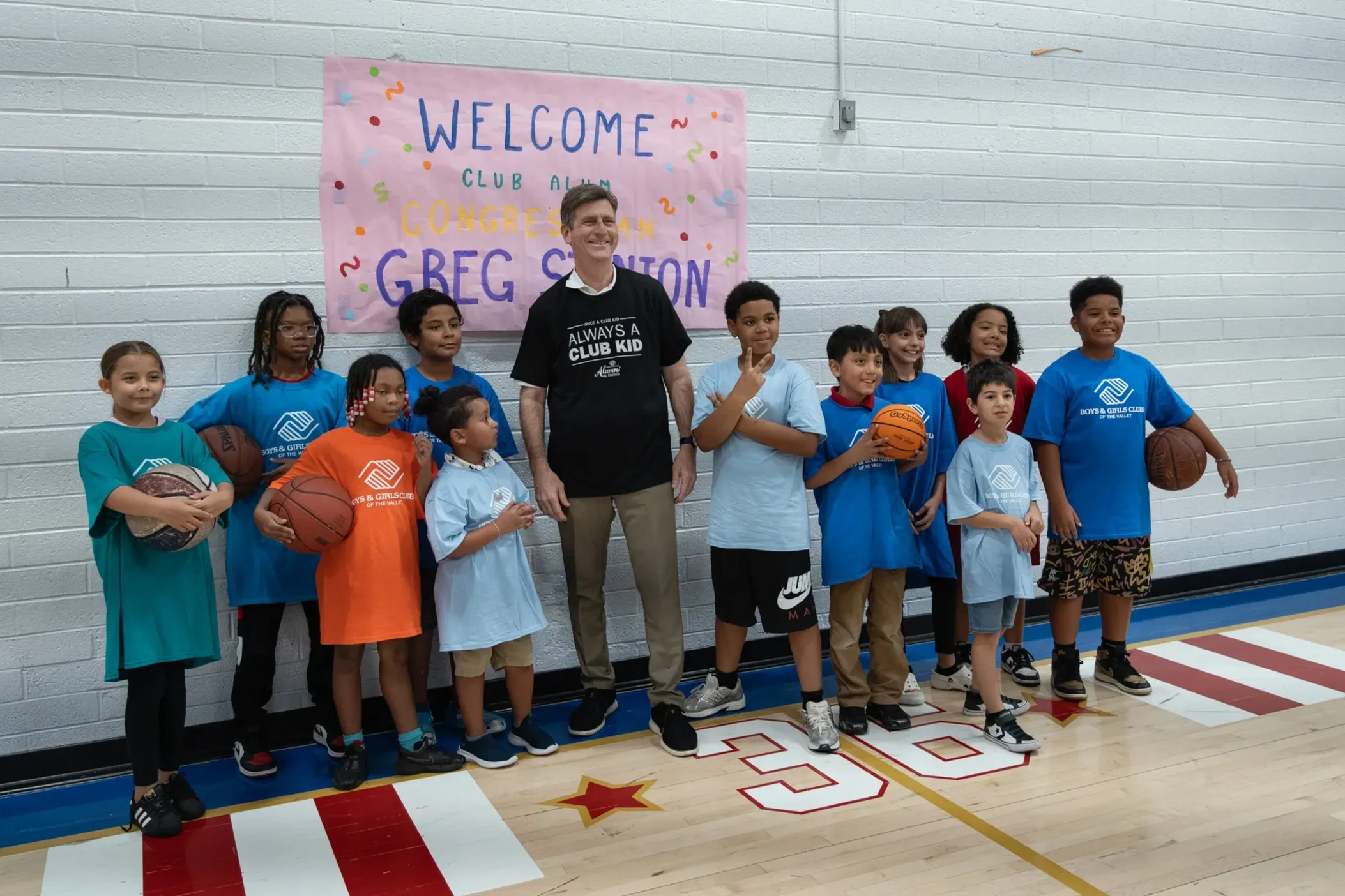 A group of children in colorful sports shirts stand on a gymnasium floor, holding basketballs, with a smiling adult man in black shirt standing behind them. A pink banner with colorful letters reading 'Welcome Club Alumni Congressional G-Beg Session' is behind them.