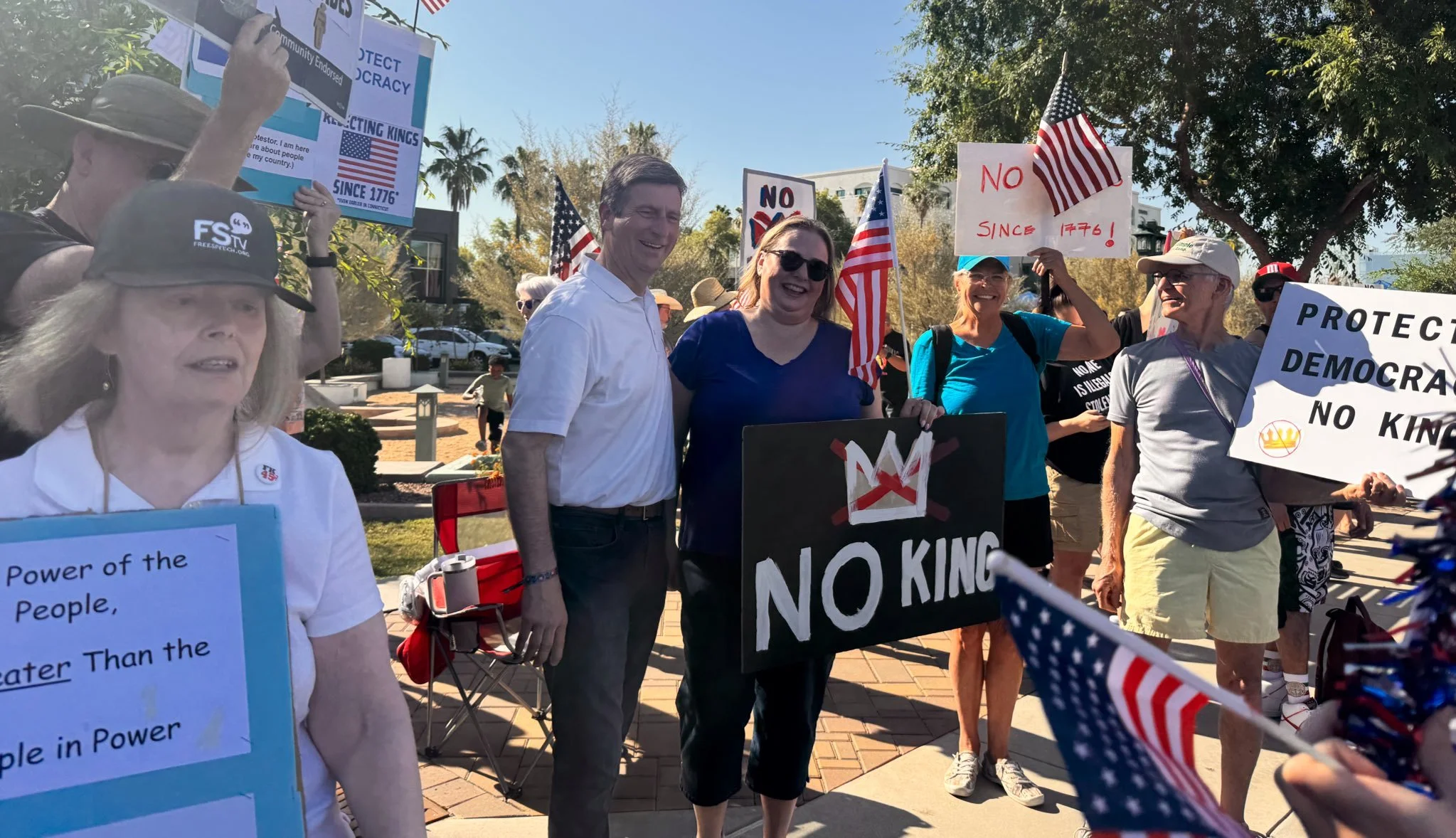 Happy protesters at a rally holding signs and American flags, with some wearing hats and sunglasses, during the day in an outdoor setting with trees and buildings in the background.