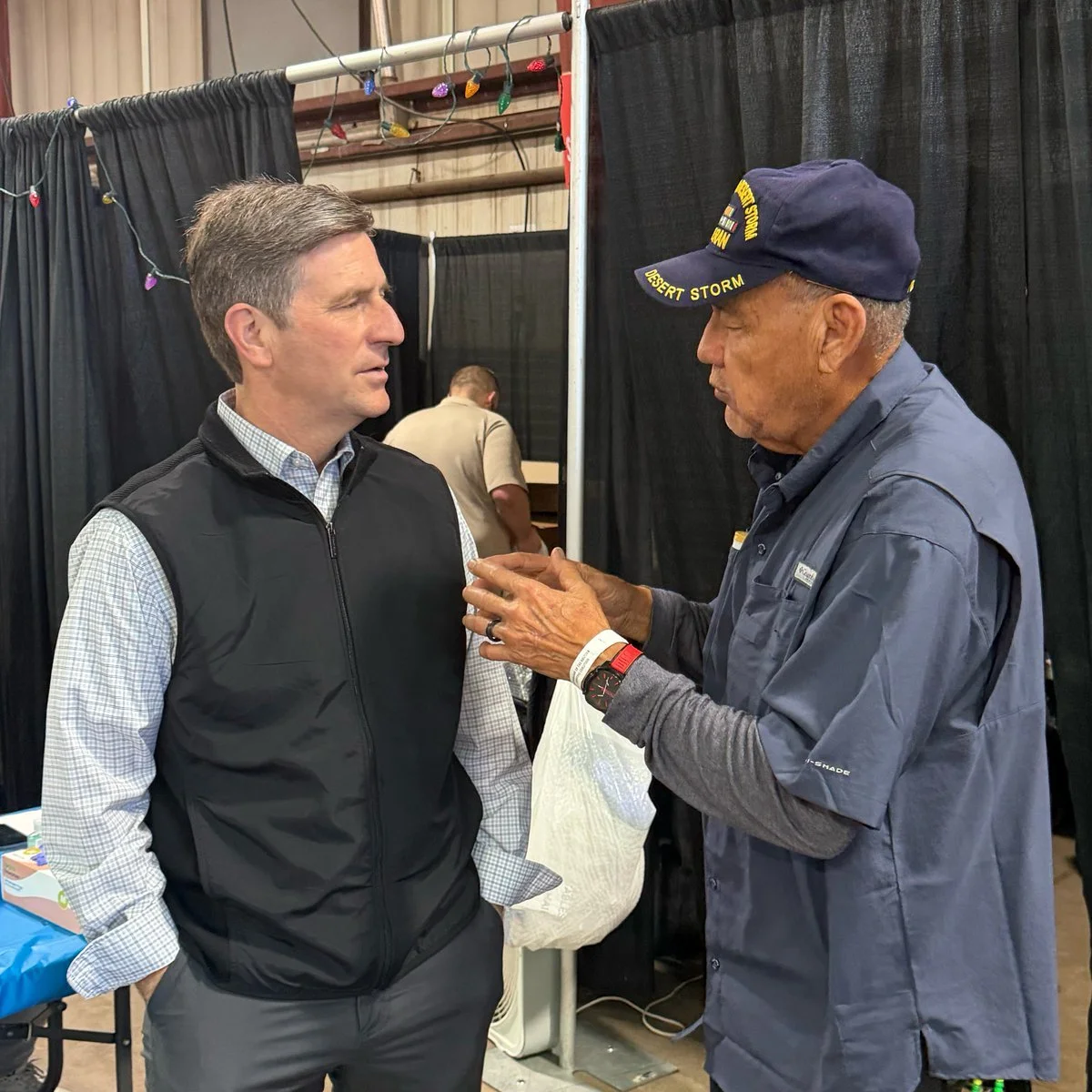 Two men are engaged in a conversation, one wearing a black vest and the other wearing a blue uniform and cap with yellow text, in an indoor setting with black curtains and fairy lights.