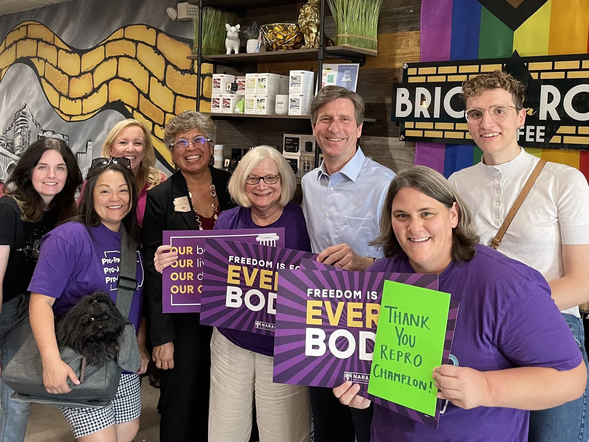 A diverse group of people gathered together, holding signs and smiling. Some signs read "FREEDOM IS EVERYBODY'S RIGHT," "THANK YOU REPRO CHAMPIONS!" and other slogans. The background features a mural with a yellow chain design and a rainbow flag.