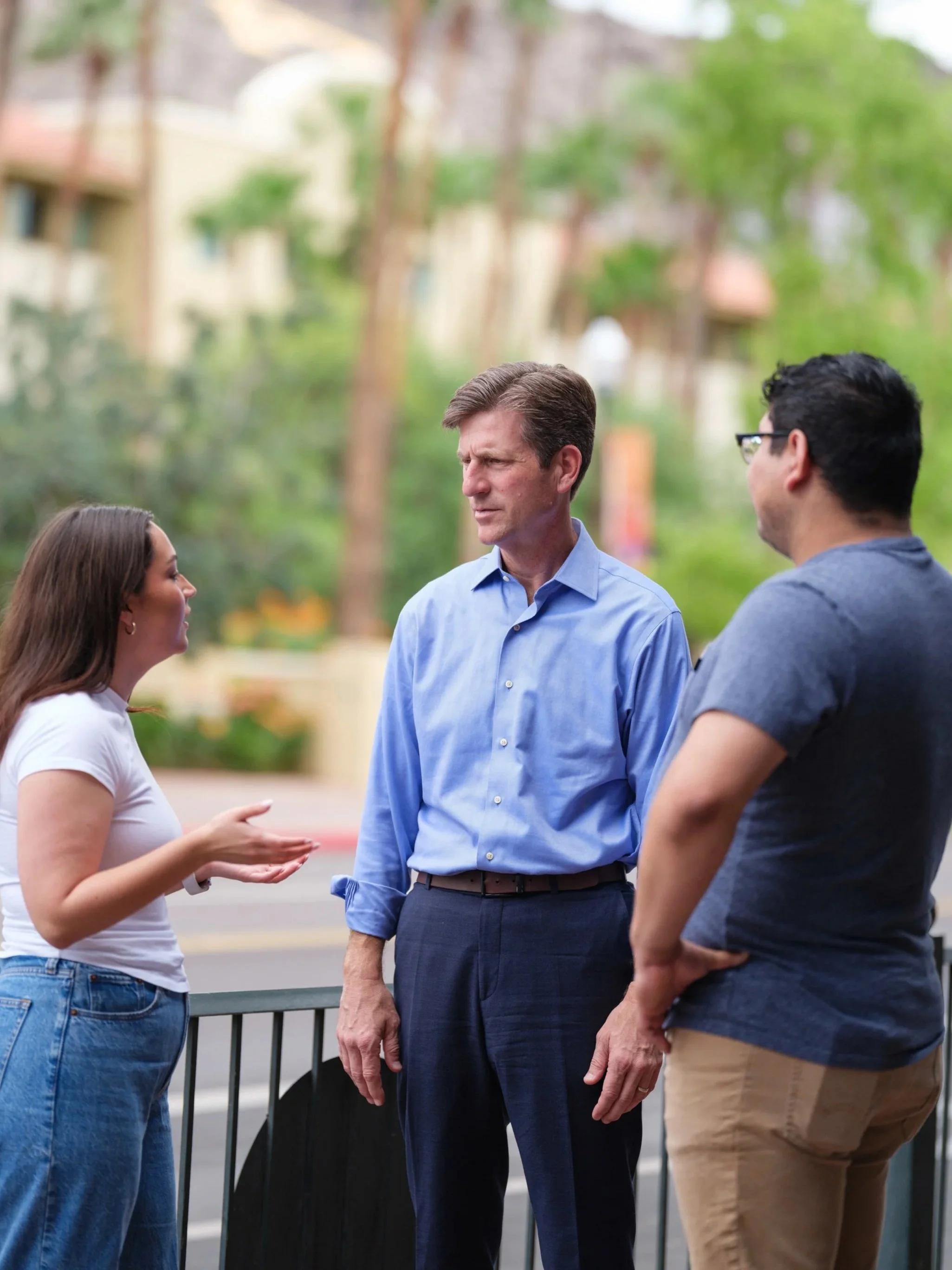 Three people engaged in conversation outdoors, with a woman on the left gesturing with her hands and two men listening, one in a light blue shirt and the other in a gray shirt, with trees and buildings in the background.