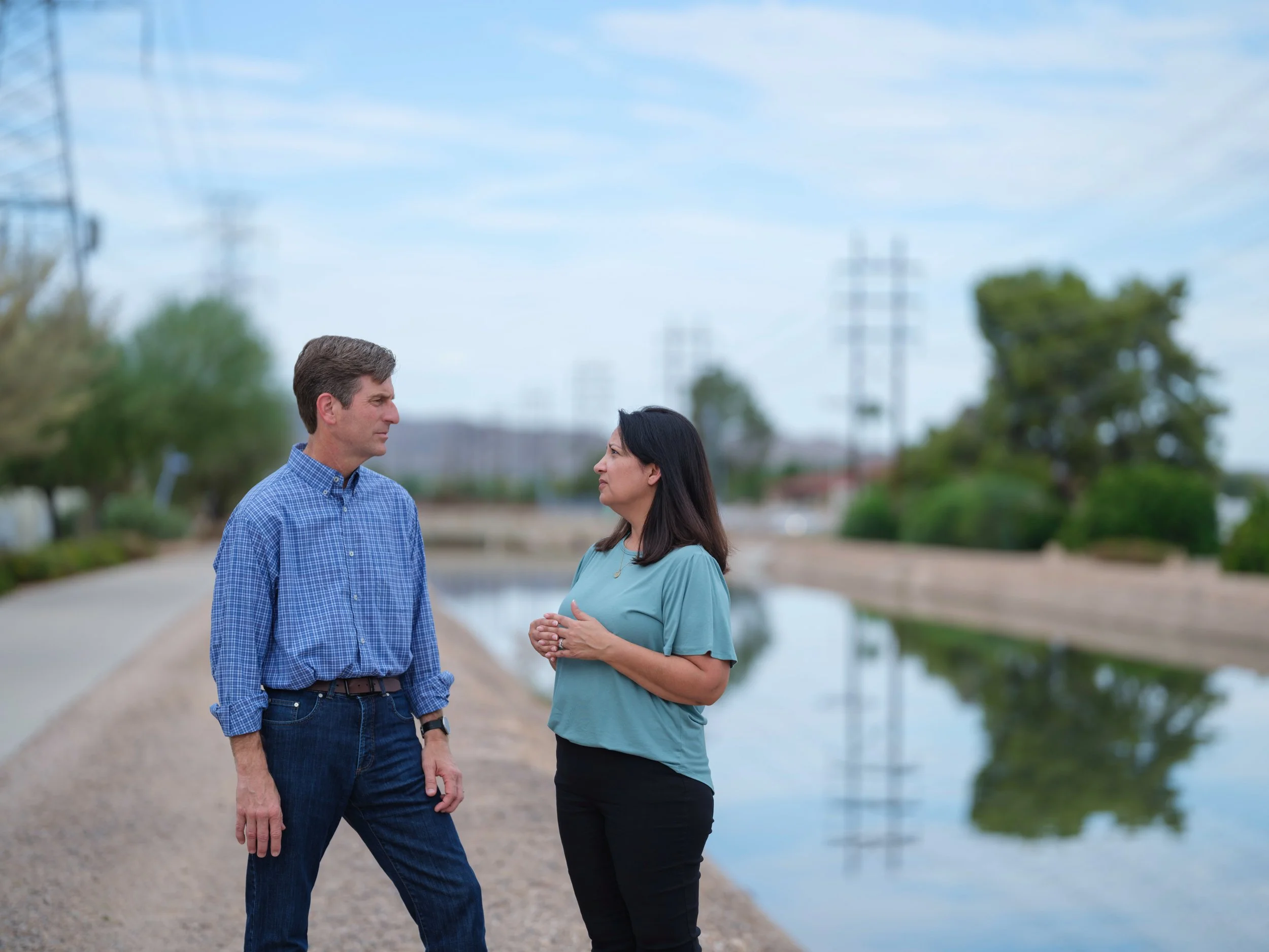 A man and a woman stand outside near a body of water, having a conversation, with power lines and trees in the background.